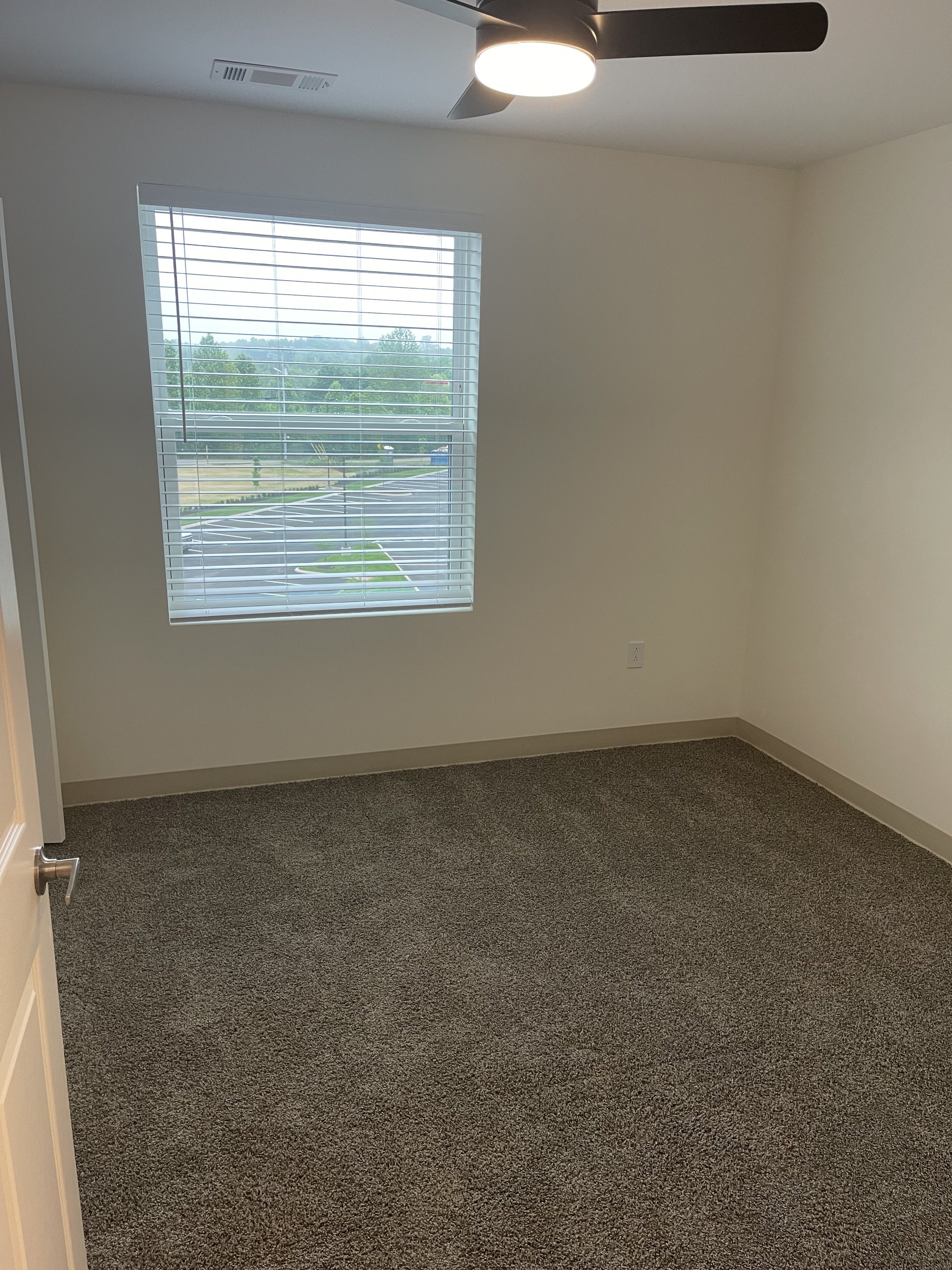 Empty room with beige walls, a window with blinds, a ceiling fan with light, and brown carpet