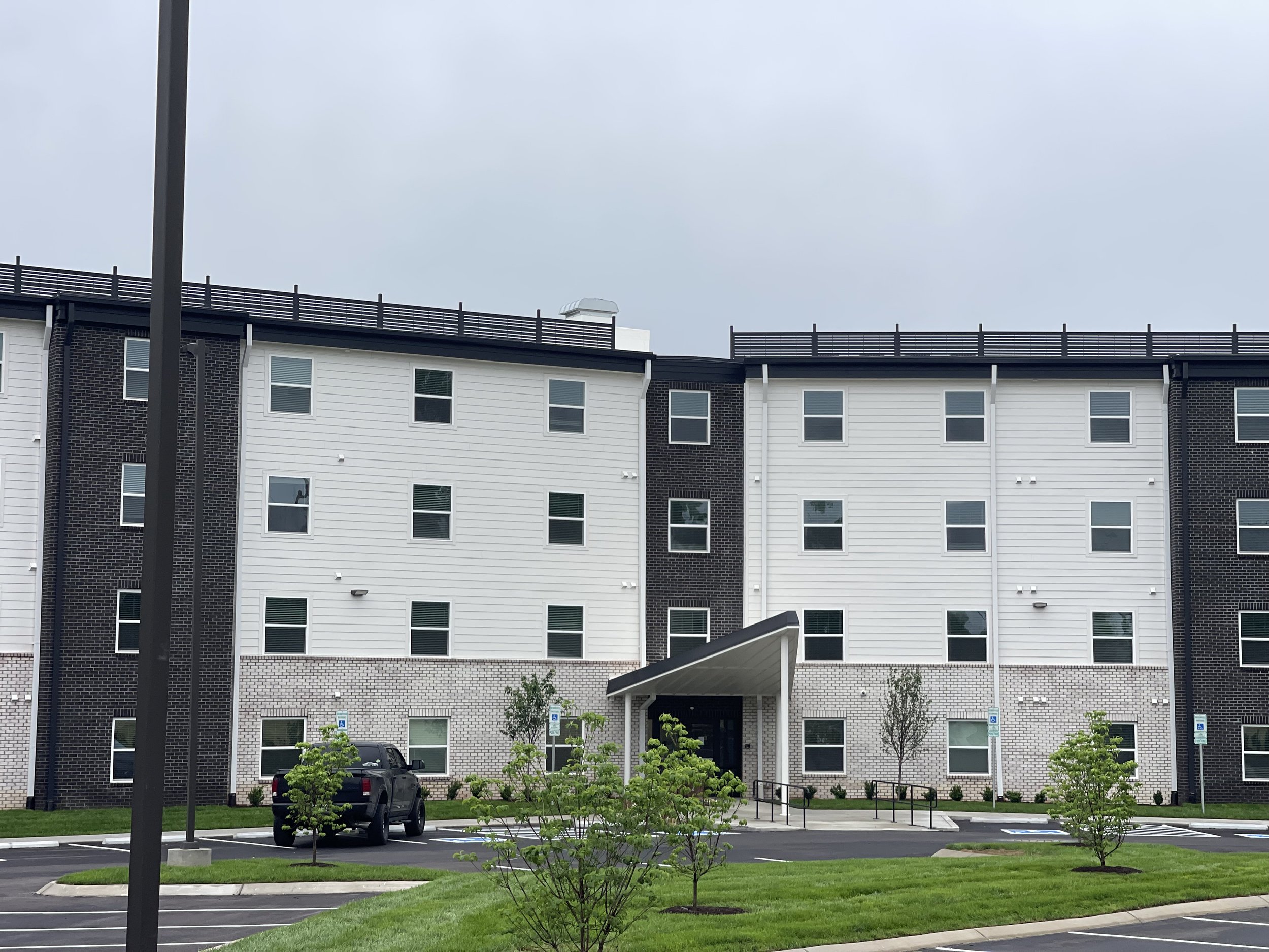 Modern multi-story apartment building with white and dark brick exterior, small trees, parked cars, and a landscaped parking lot.