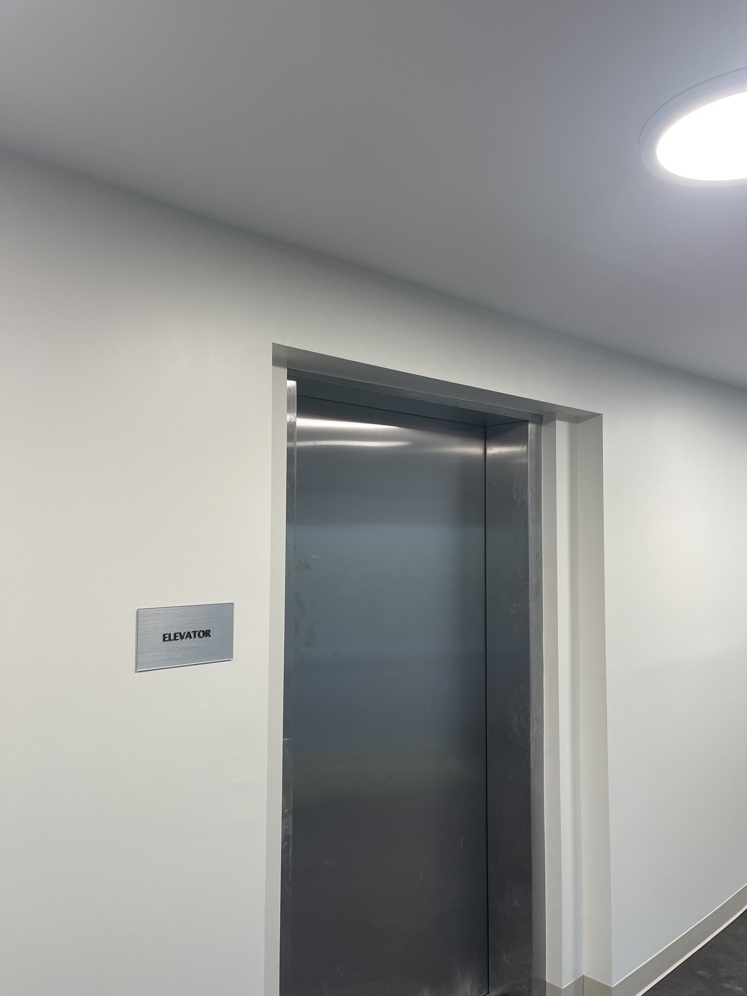 Stainless steel elevator door in a white wall, with a sign on the wall reading 'ELEVATOR'.