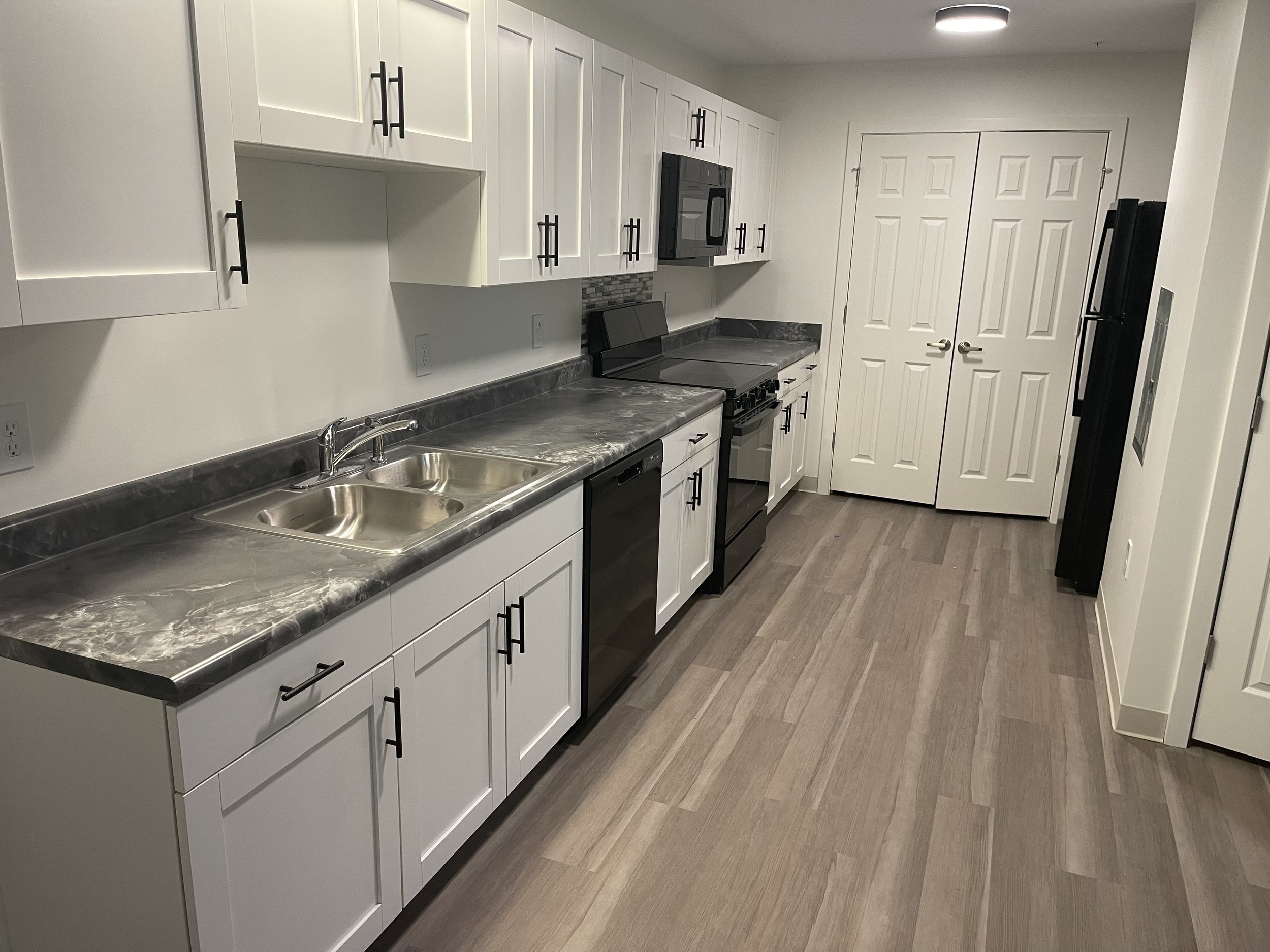 Modern kitchen with white cabinets, black hardware, dark marble countertop, black appliances, double sink, and light wood flooring.