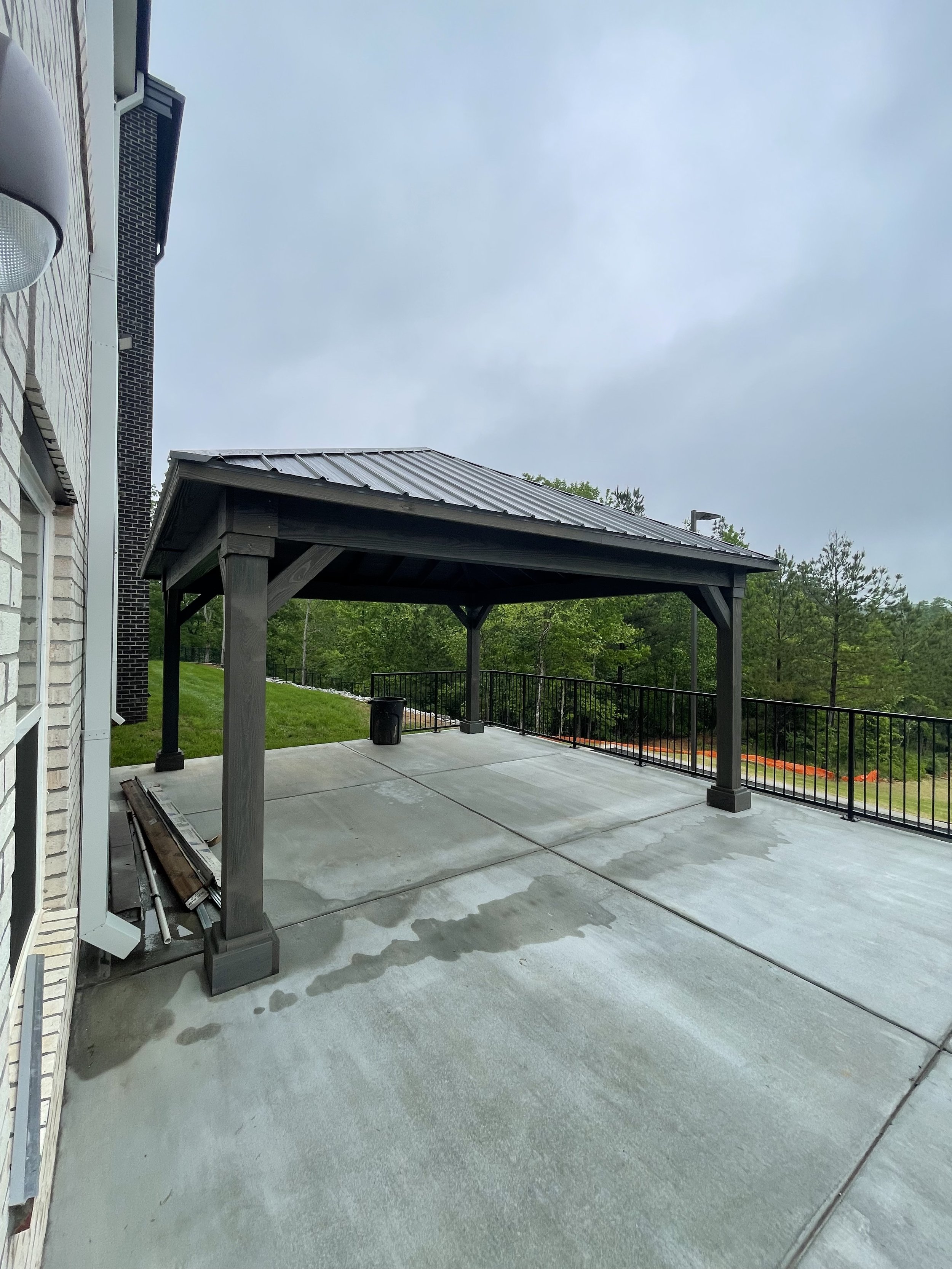 Empty outdoor patio with a covered pavilion, concrete flooring, black metal railing, a trash can, and trees in the background overcast sky