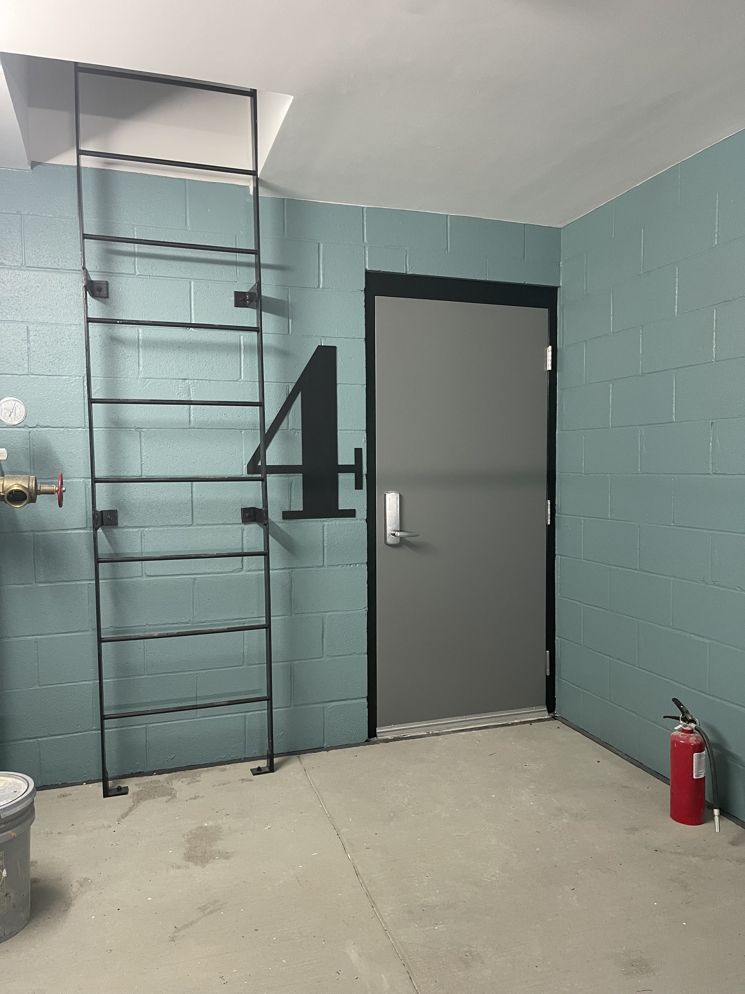 A door marked with the number four, with a black metal ladder mounted on a teal cinder block wall to its left, and a red fire extinguisher on the floor to its right.