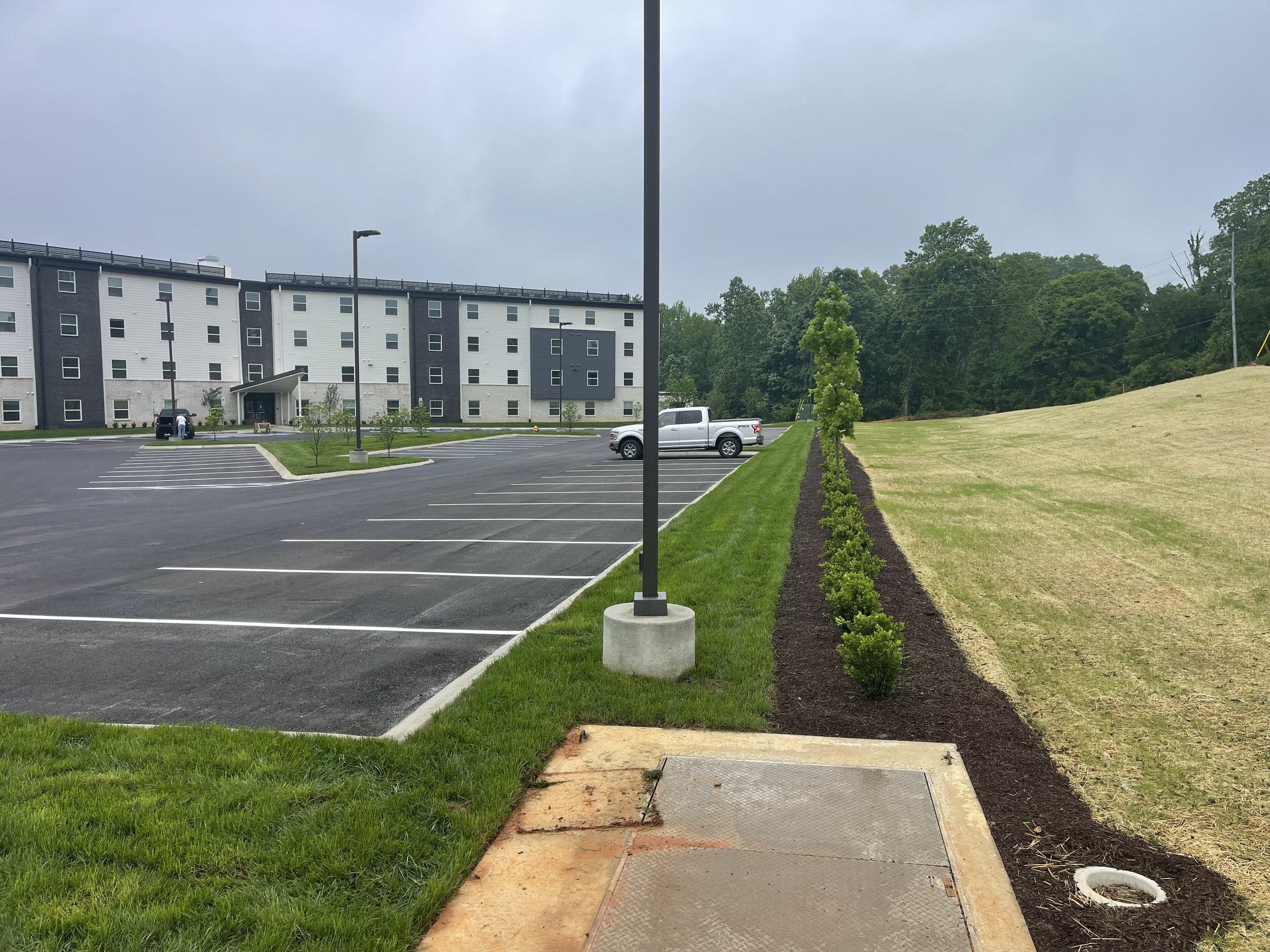 Empty parking lot near modern multi-story apartment building with new trees and grass landscaping, cloudy sky.