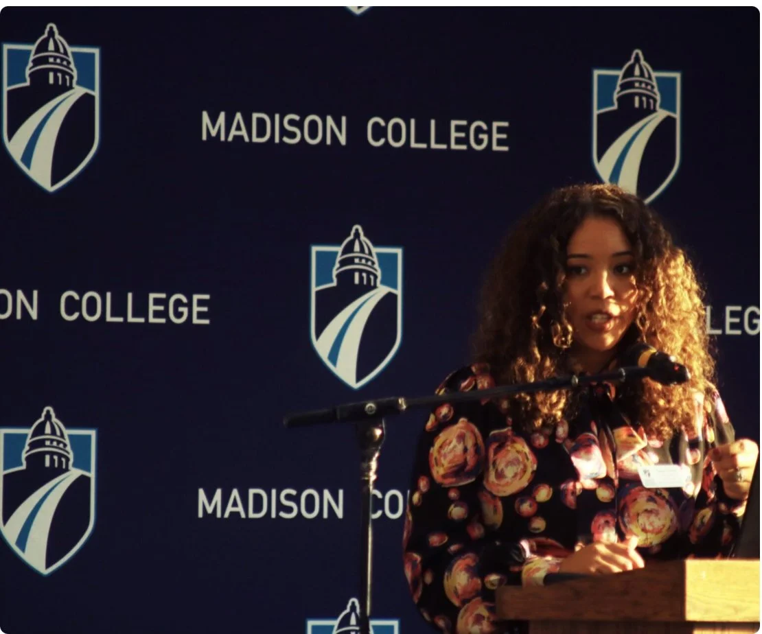 A woman with curly hair speaking at a podium with a Madison College backdrop behind her.