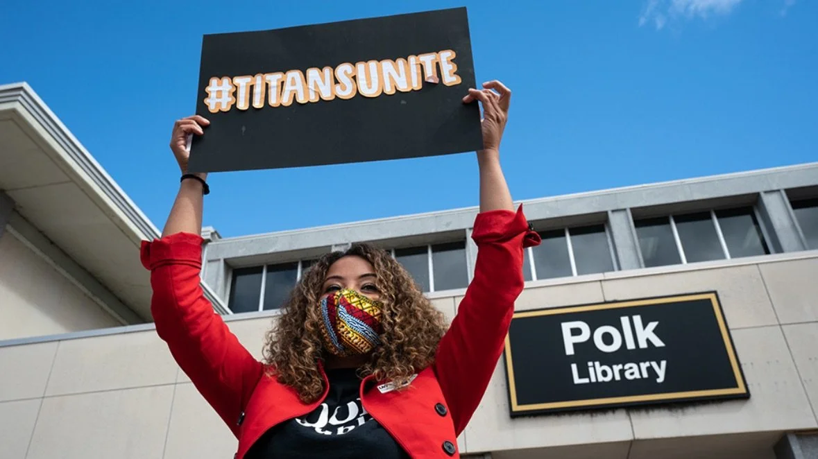 A woman wearing a colorful face mask and red jacket holding a sign that says #TITANSUNITE outside Polk Library.