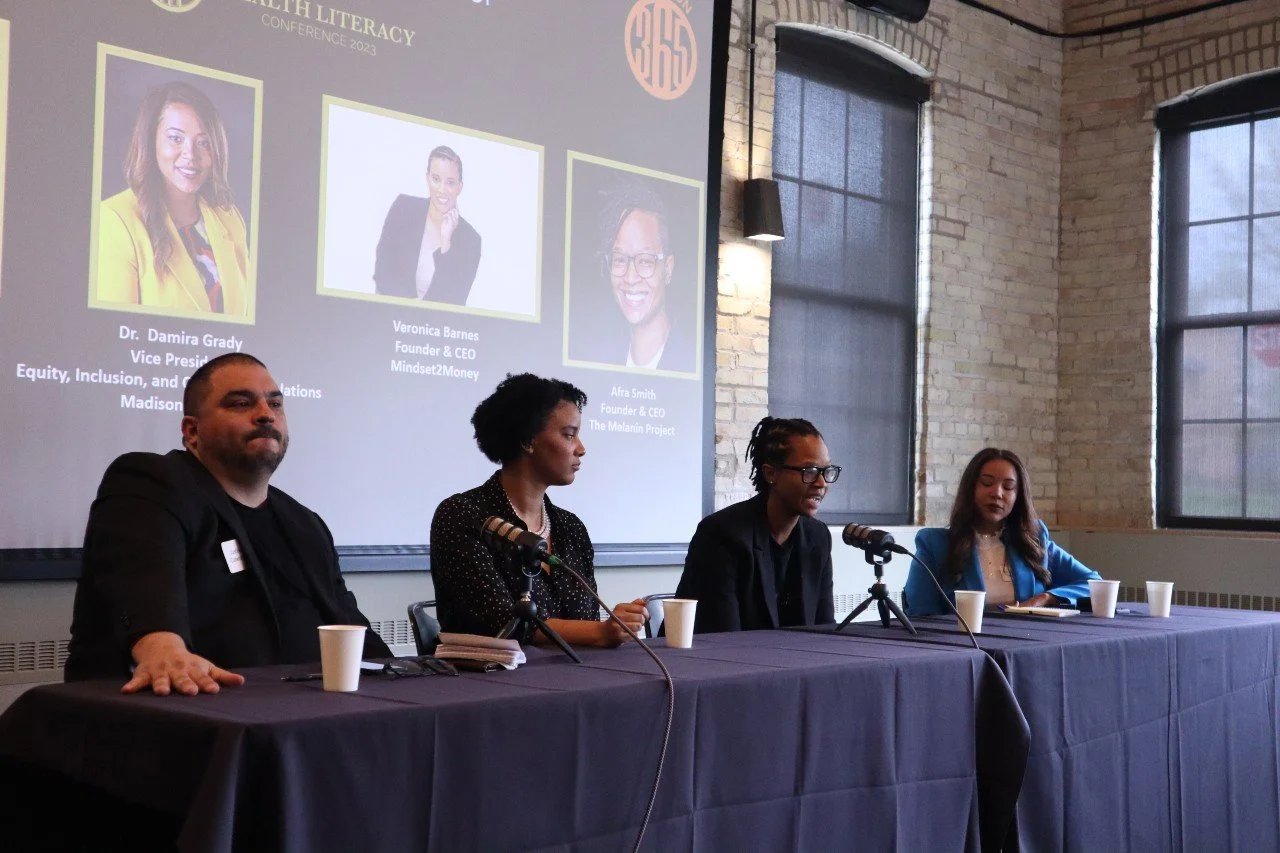 Panel of four diverse individuals sitting at a table with microphones during a conference, with a large screen behind them displaying photos and names of speakers.