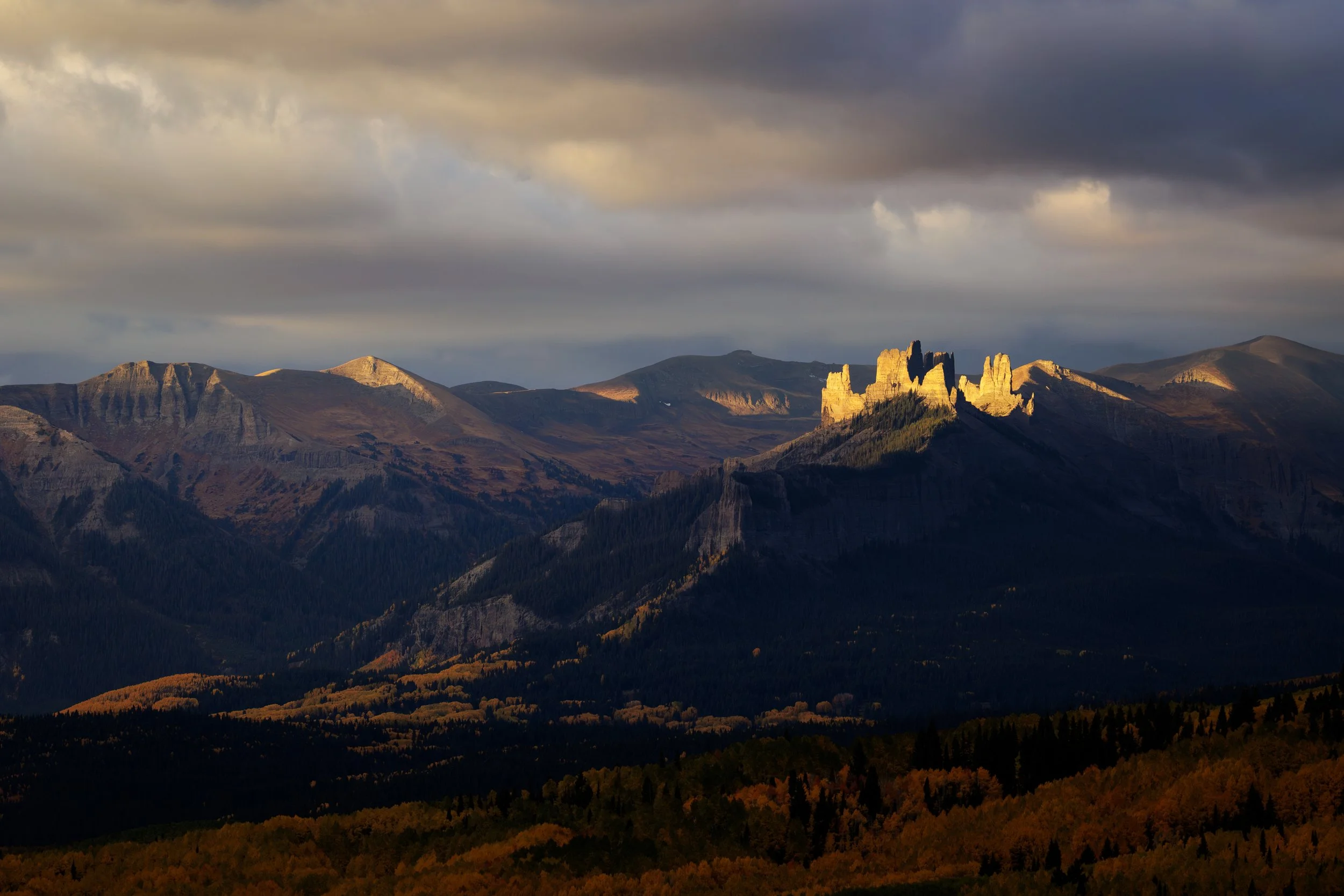 Sunlit rock formations on a mountain ridge with dark clouds overhead and a forested landscape below.