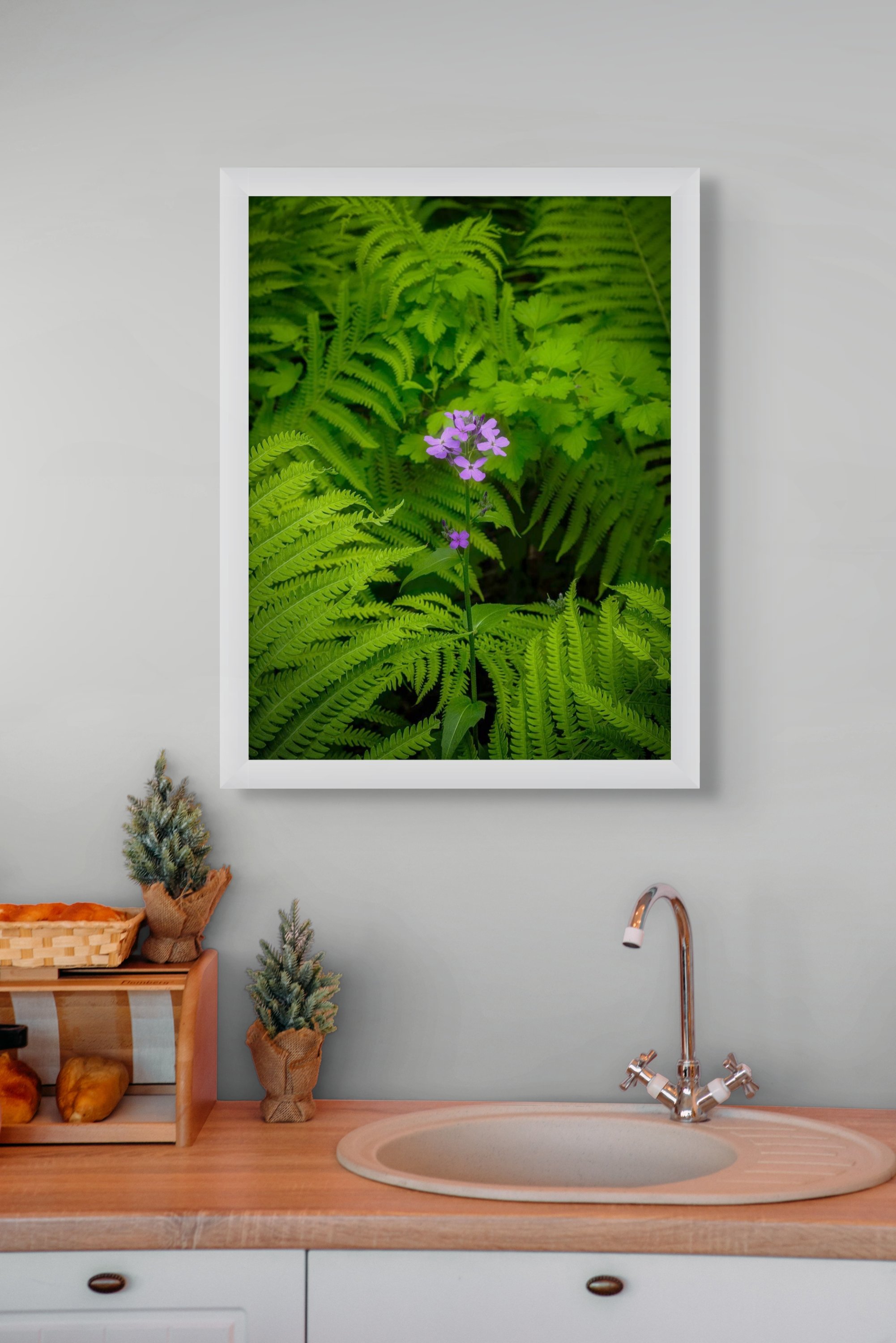 A framed photograph of a purple flower surrounded by green ferns and foliage hangs above a kitchen sink with a chrome faucet. The kitchen has a wooden countertop and small potted plants on the counter.