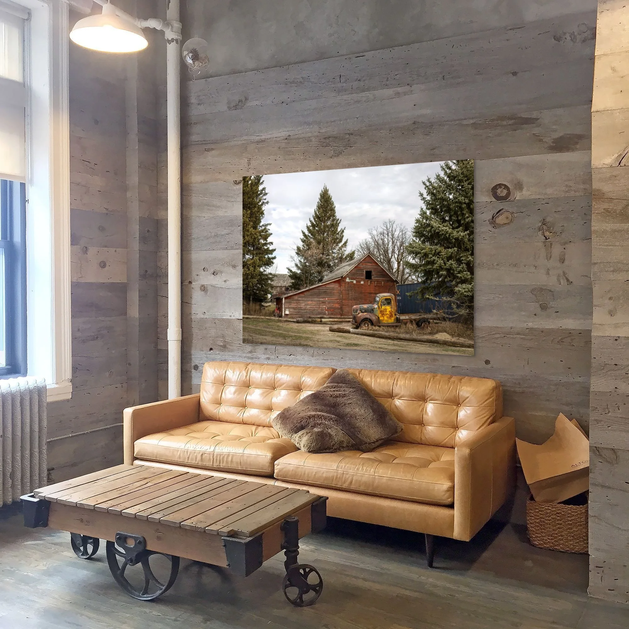 Living room with a tan leather couch, a faux fur pillow, a wooden coffee table on wheels, a window with blinds, and a large window showing a rural scene with a barn, an old truck, and trees.