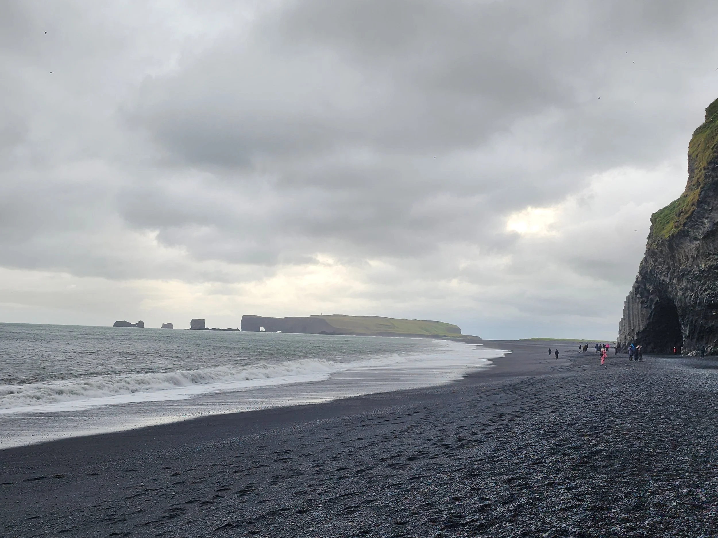 A black sand beach with people walking near a large rocky cliff and distant islands under a cloudy sky.