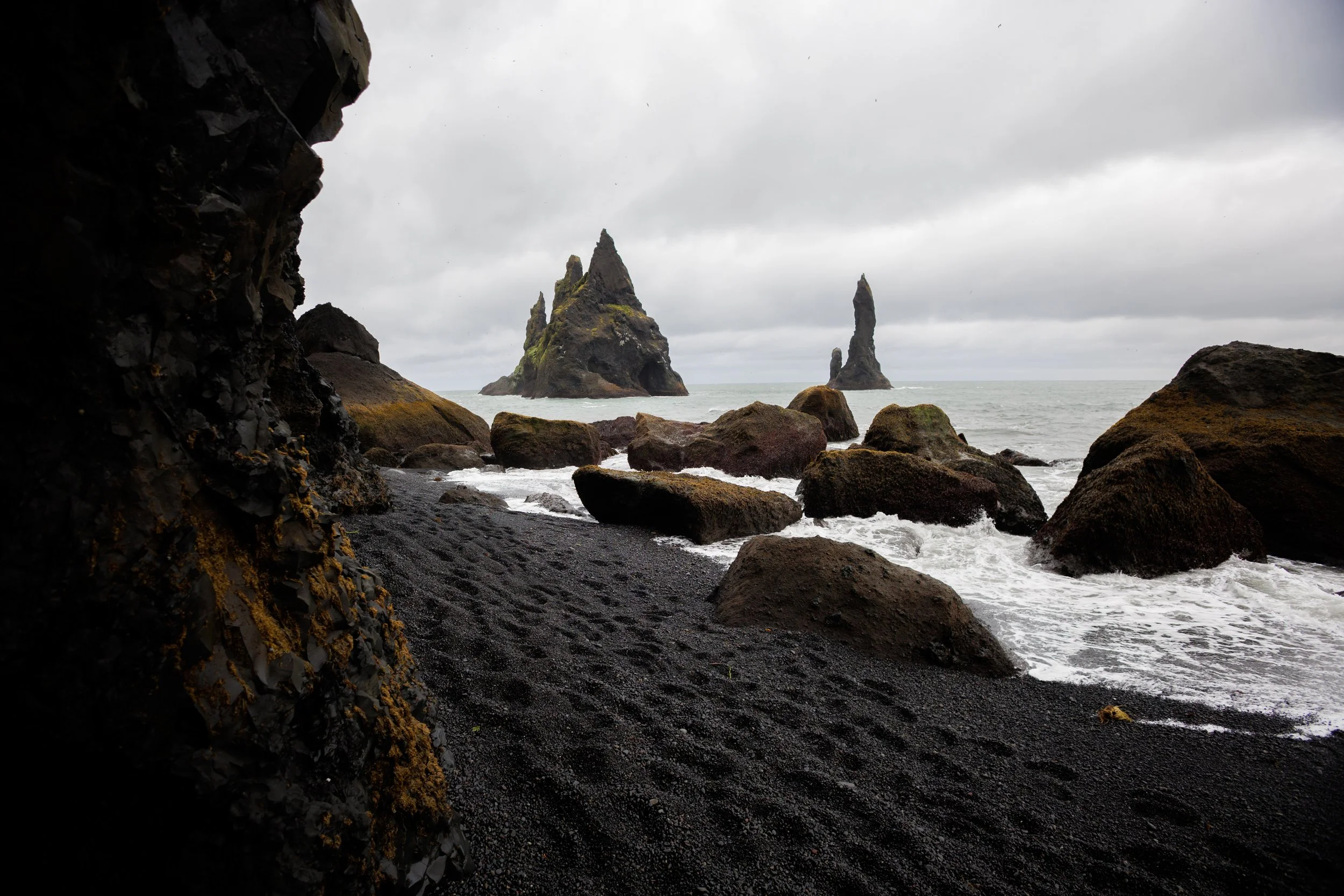 A view from inside a dark cave looking out to a black sand beach with large rocks and sea stacks in the water under a cloudy sky.