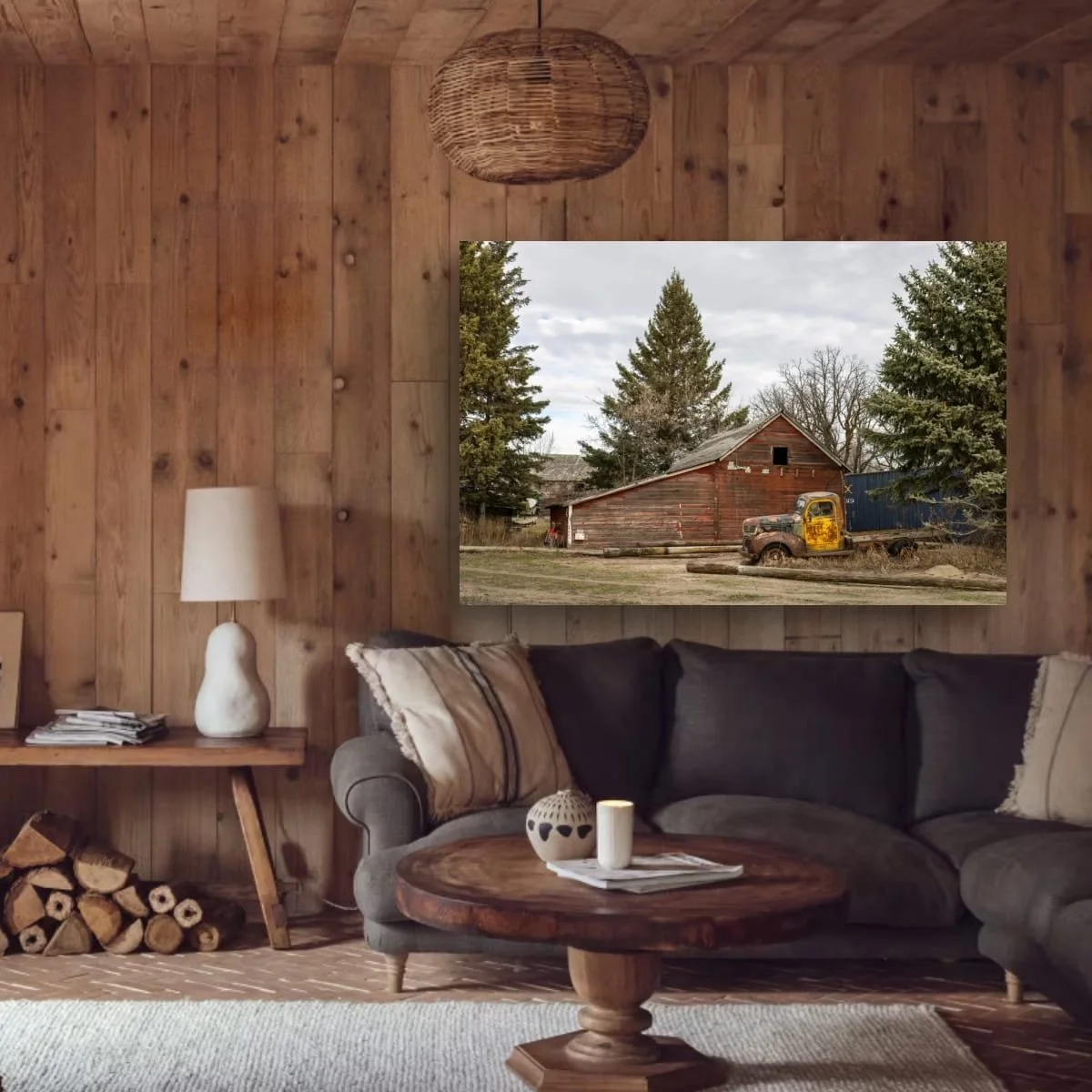 Living room with wood-paneled walls, a gray sofa with pillows, a round wooden coffee table with candles and a magazine, a side table with a white lamp and magazines, a stack of firewood, and a large window showing an outdoor scene with trees, an old 