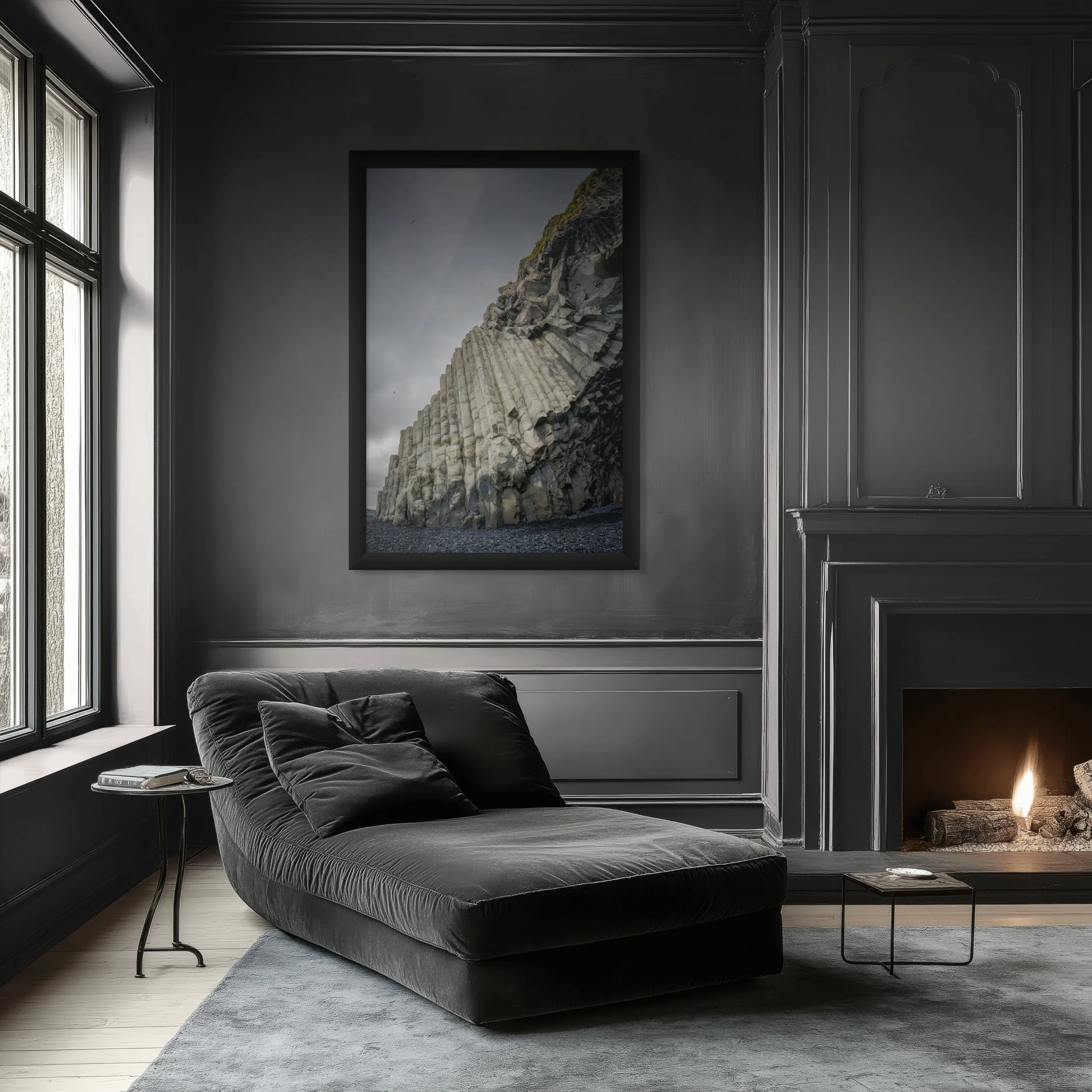 A modern living room with dark gray walls, black framed artwork of a rocky cliff, a black sofa with black cushions, a small side table with a telephone, a fireplace with logs and a small table nearby, and a large window letting in natural light.