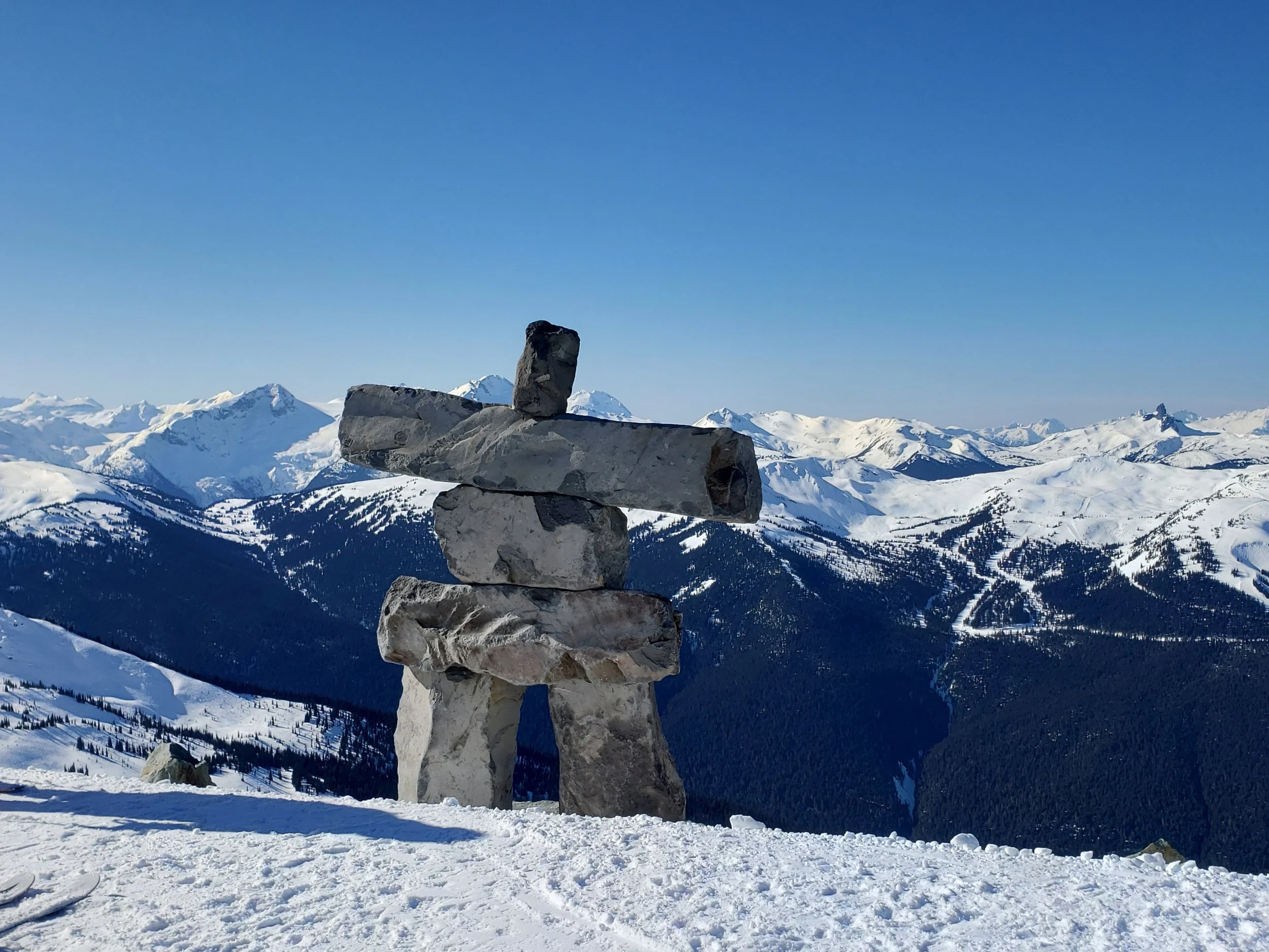 A stone cairn on a snowy mountain summit with snow-capped mountains in the background under a clear blue sky.