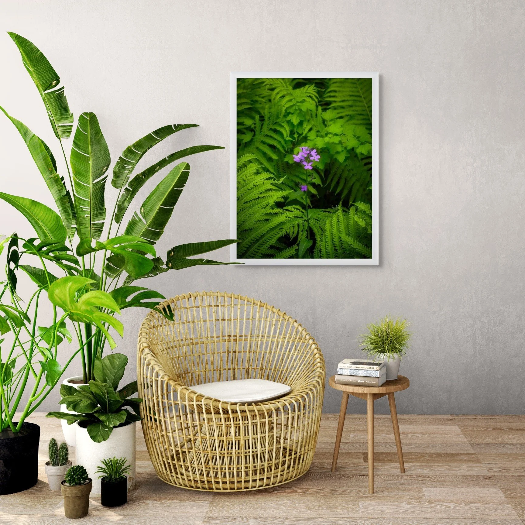 A cozy living room corner with a large green potted plant, a rattan armchair with a cushion, a small wooden side table with books and a small potted plant, and a framed picture of lush green ferns and purple flowers on the wall.