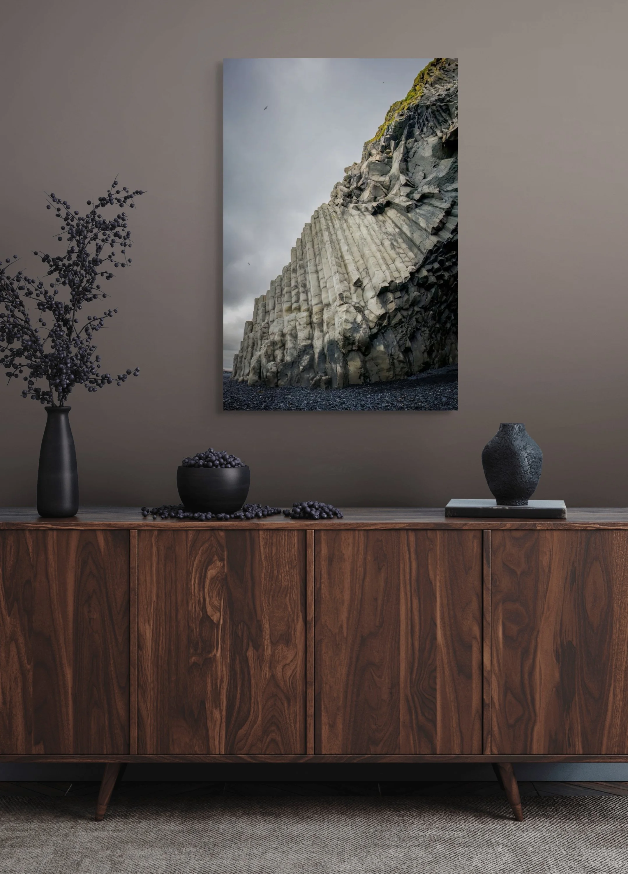A photograph of a tall, rocky cliff with vertical grooves under a cloudy sky, hung on a beige wall above a mid-century modern wooden sideboard. The sideboard holds a tall black vase with purple berries, a black bowl filled with purple berries, and a 