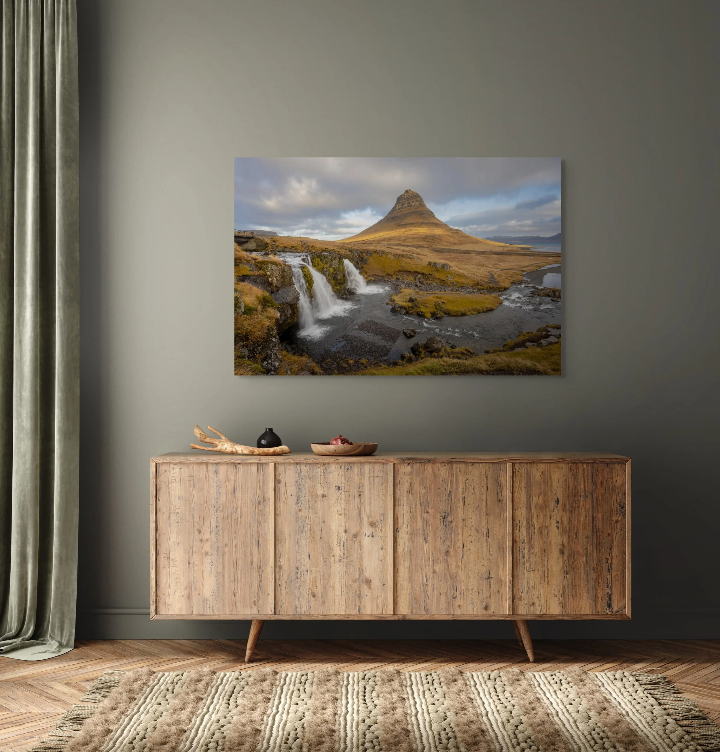 Living room with a large landscape photograph of Kirkjufell mountain and waterfalls on a gray wall, a wooden sideboard, a black vase, a wooden bowl, and a beige patterned rug.