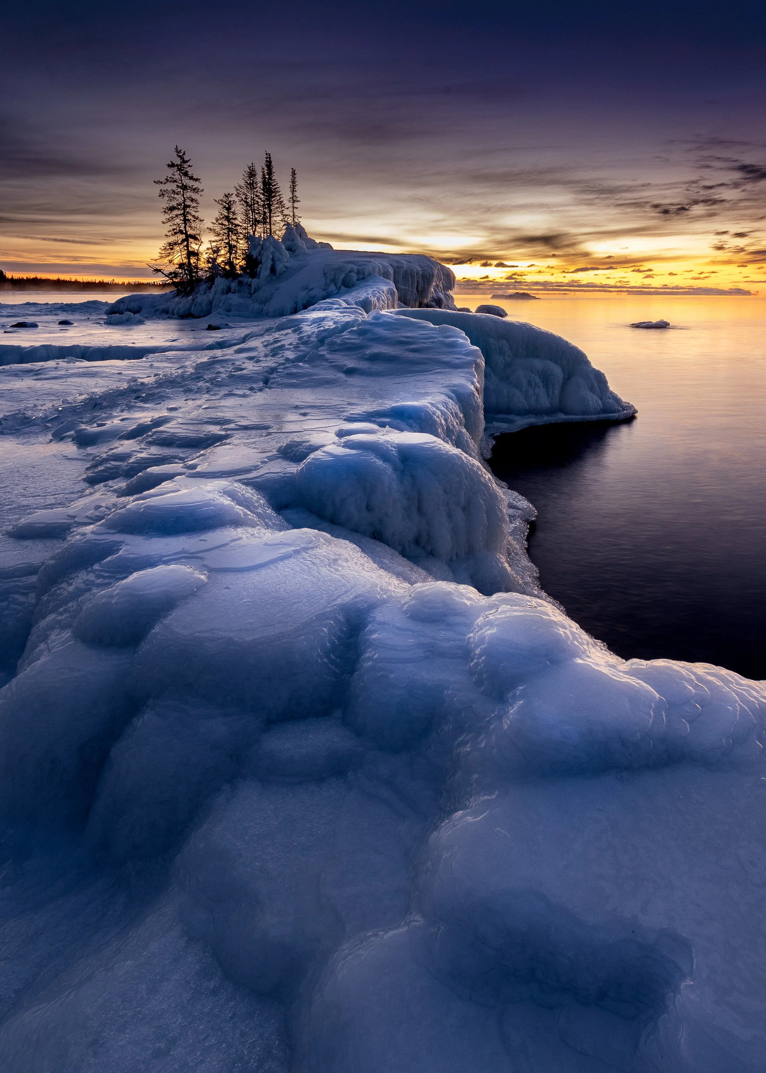 A rocky point covered in wind-sculpted ice and snow extends into a calm lake beneath a purple and orange sunrise sky, with a small cluster of snow-covered evergreen trees at its tip and a faint shoreline visible in the distance.