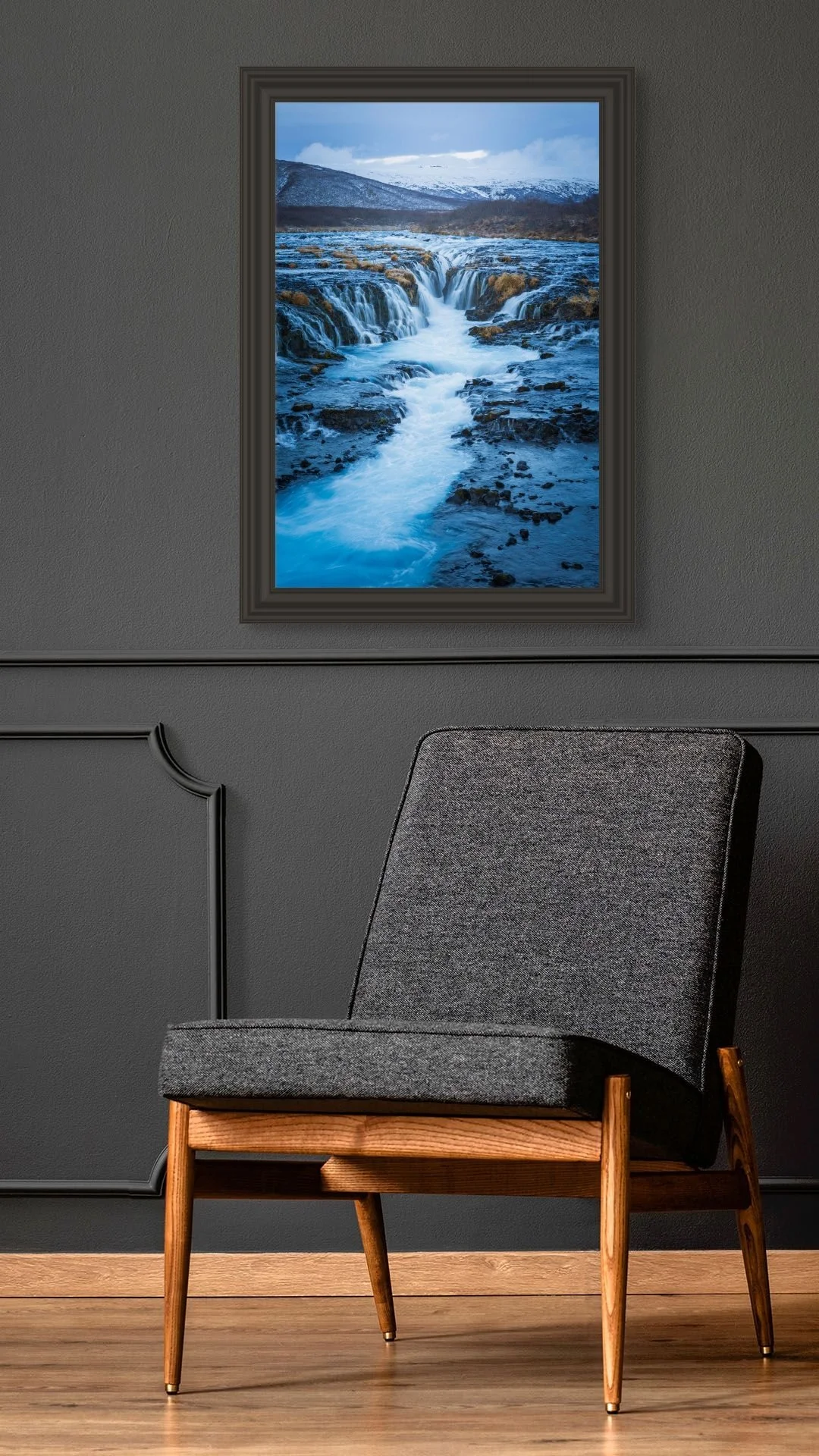 A modern gray chair with wooden legs in front of a dark gray wall. A framed photograph of a cascading mountain stream with snow-capped mountains in the background hangs on the wall.