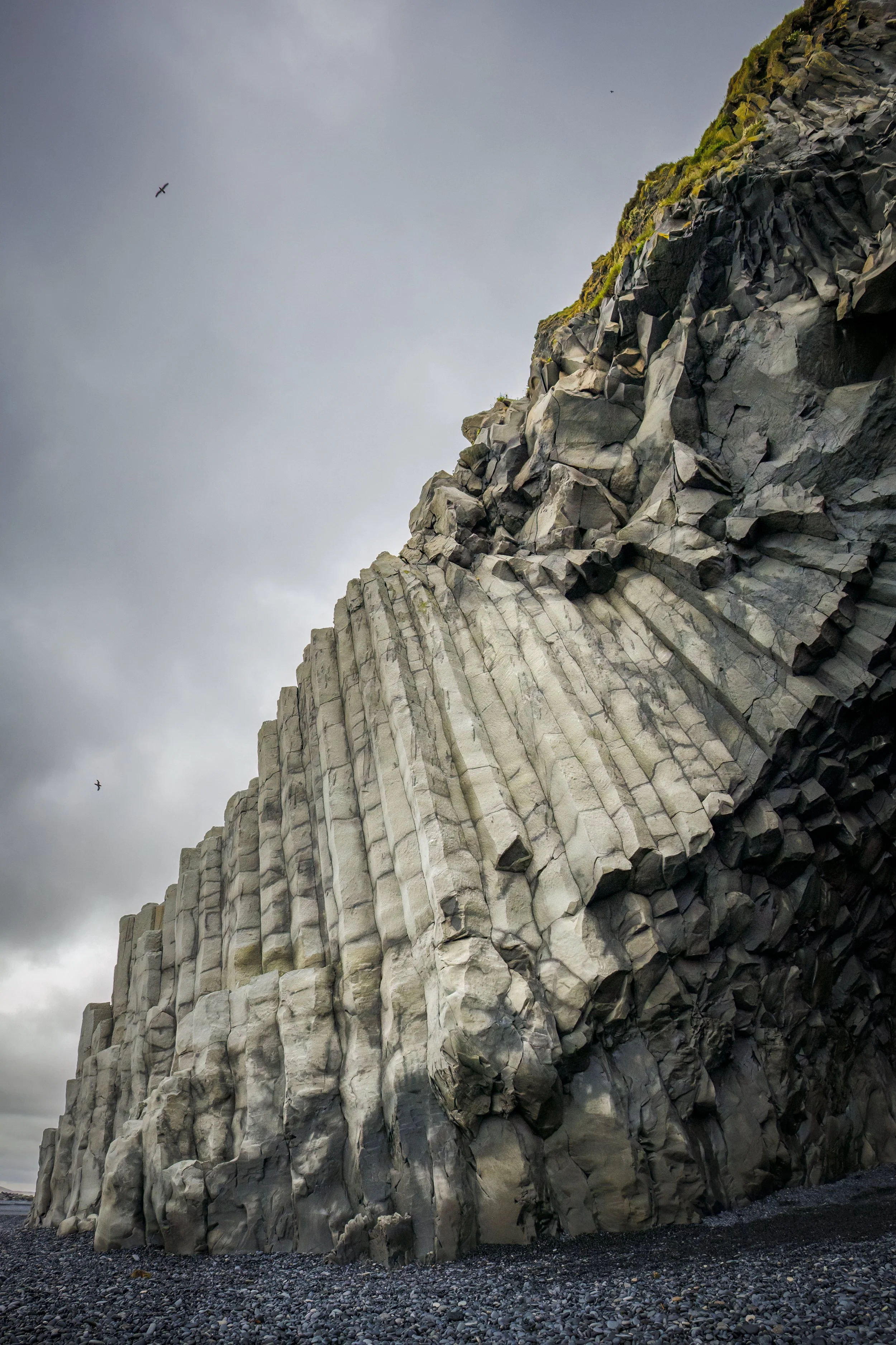 Cliffs with distinct hexagonal basalt columns on rocky shoreline under cloudy sky, birds flying overhead.