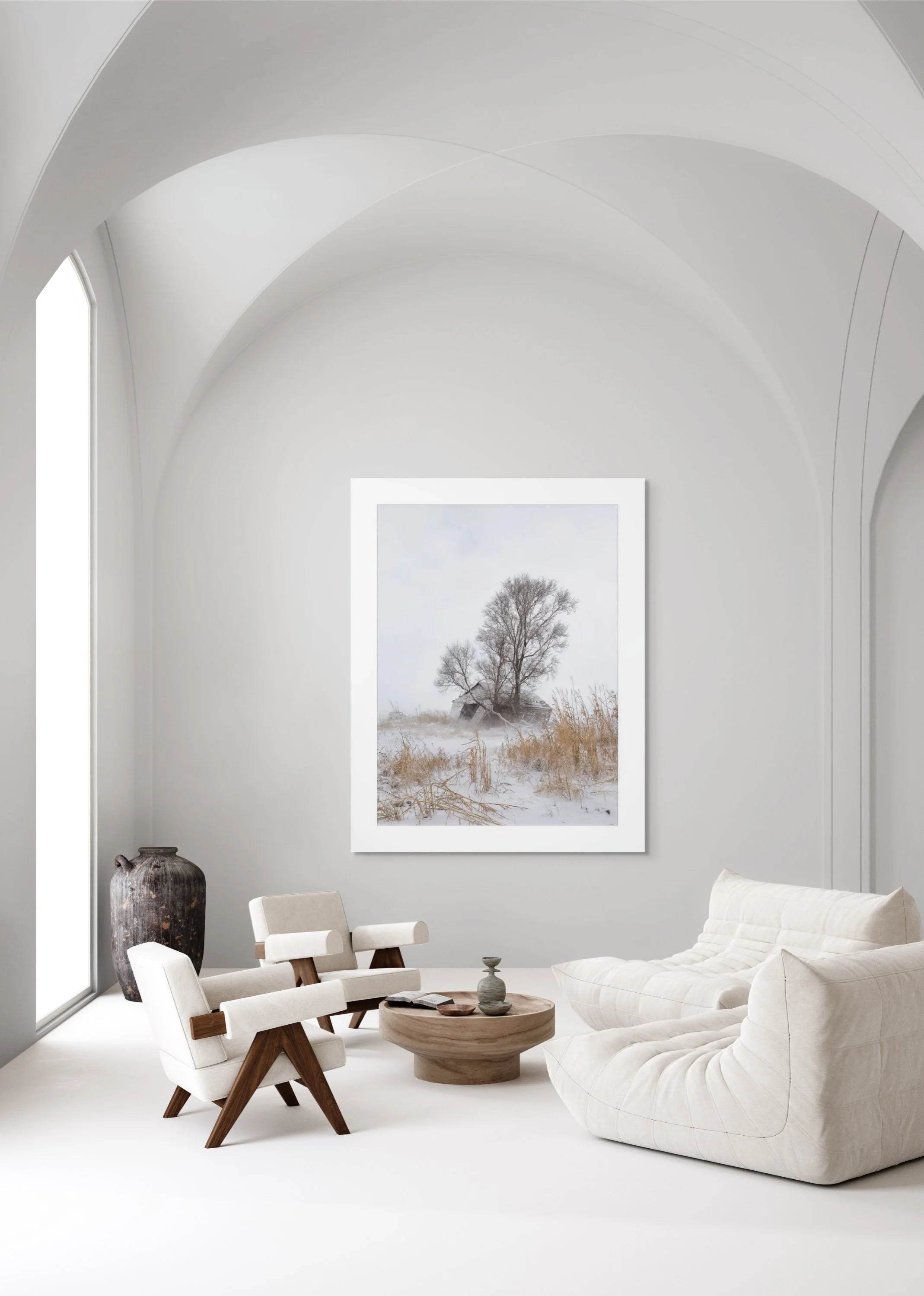 Minimalist living room with white walls, arched ceiling, large window, a white sofa, two white chairs, a wooden coffee table, a large vase, and landscape painting of a winter scene.