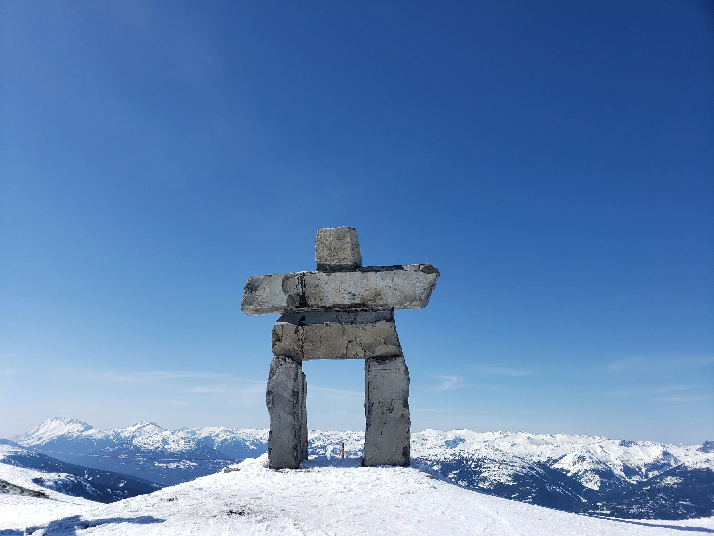 A stone monument resembling a figure with a rectangular head, arms, and legs, standing on a snowy mountain peak with snow-covered mountains and clear blue sky in the background.