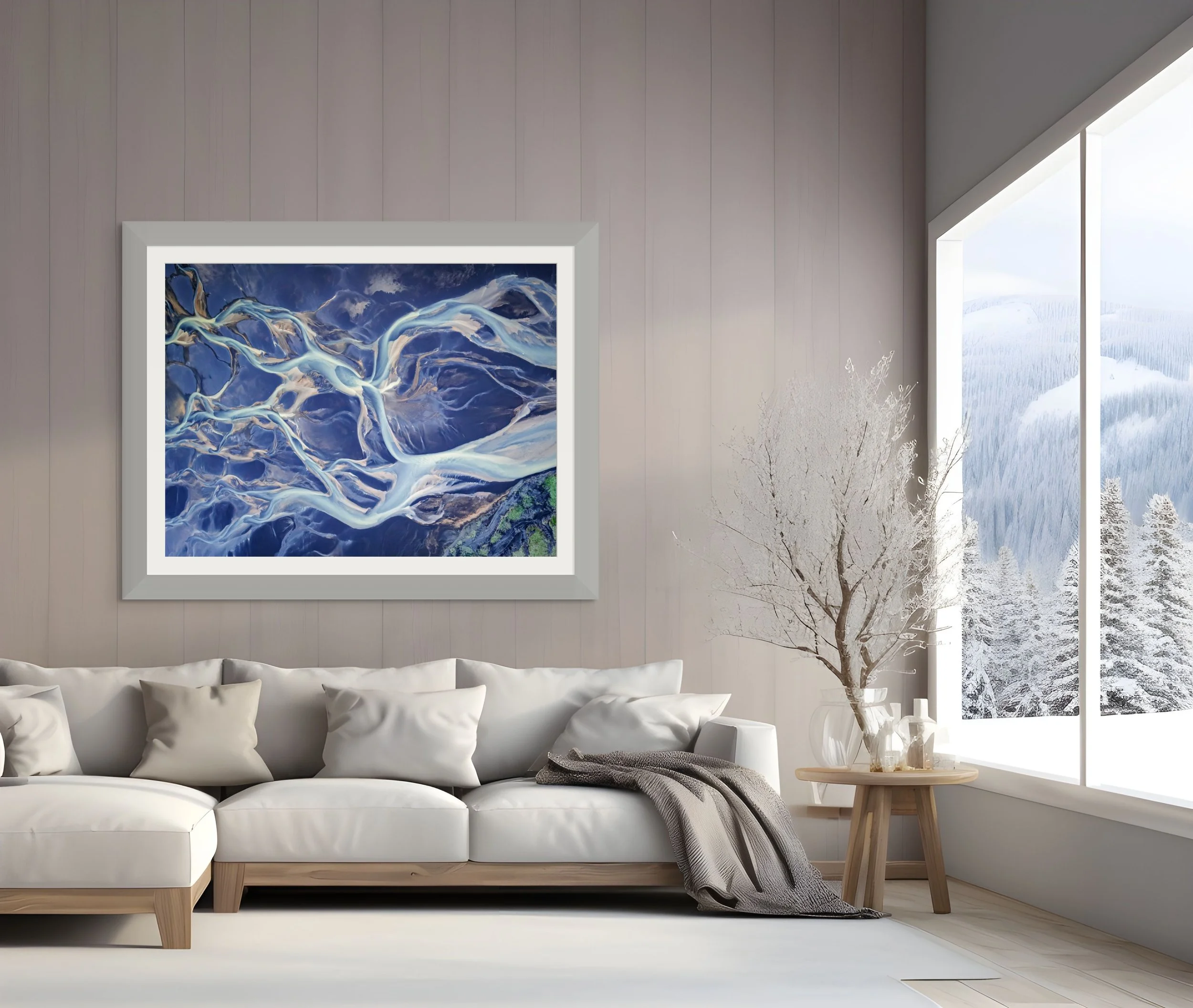 A modern living room with a white sofa, a framed abstract art piece on a light-colored wall, a small wooden table with a glass vase and winter branches, and large windows showing snowy mountains and trees outside.