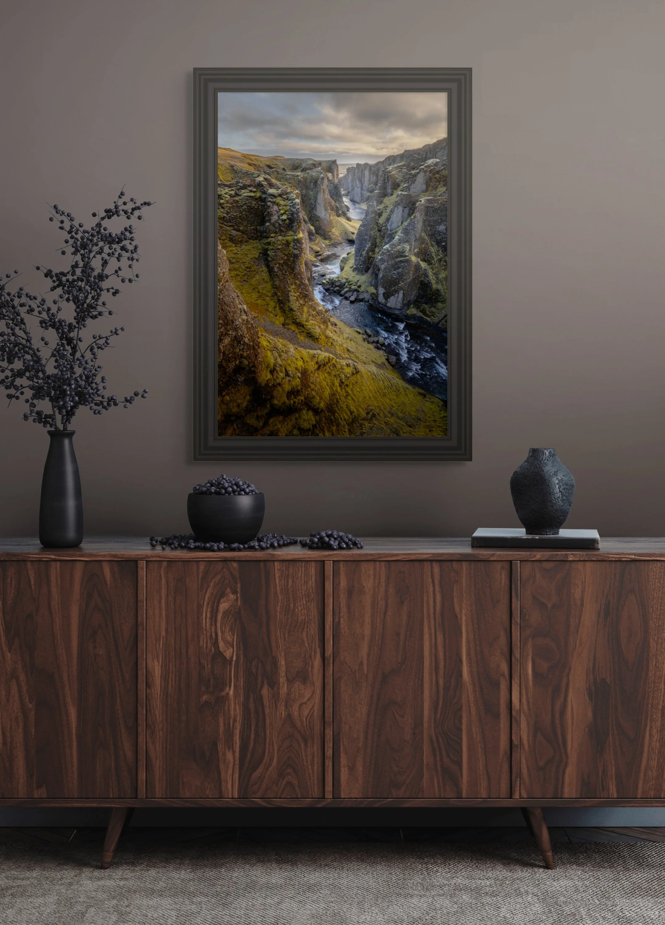 A framed photograph of a river canyon landscape with moss-covered rocks and cloudy sky, hanging on a beige wall above a wooden sideboard decorated with black vases and a bowl of berries.