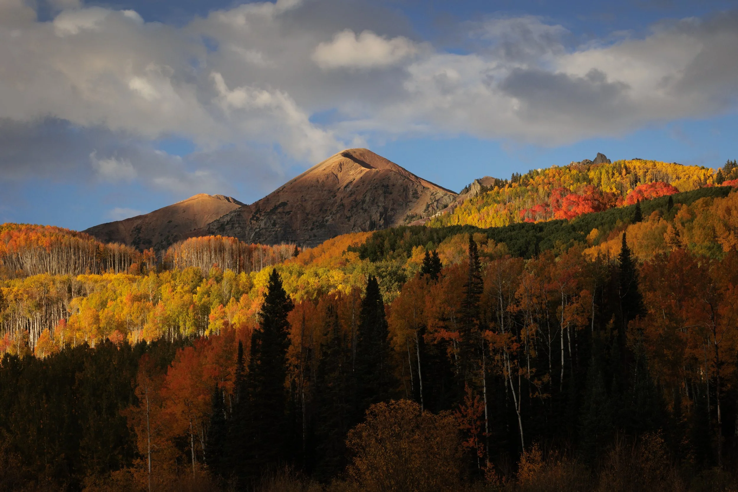 A mountain landscape during autumn with colorful fall foliage, dark evergreen trees in the foreground, rolling hills, and a partly cloudy sky.