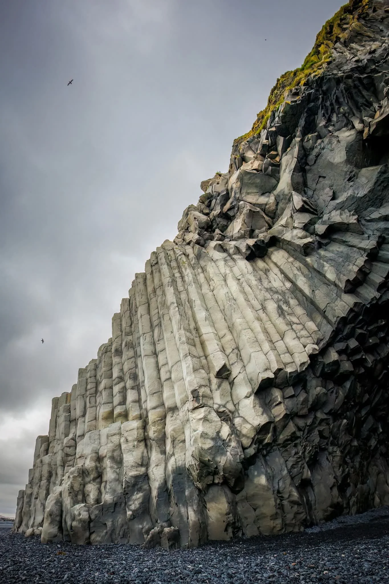 View of a towering cliff made of hexagonal basalt columns, with two birds flying in cloudy sky above.