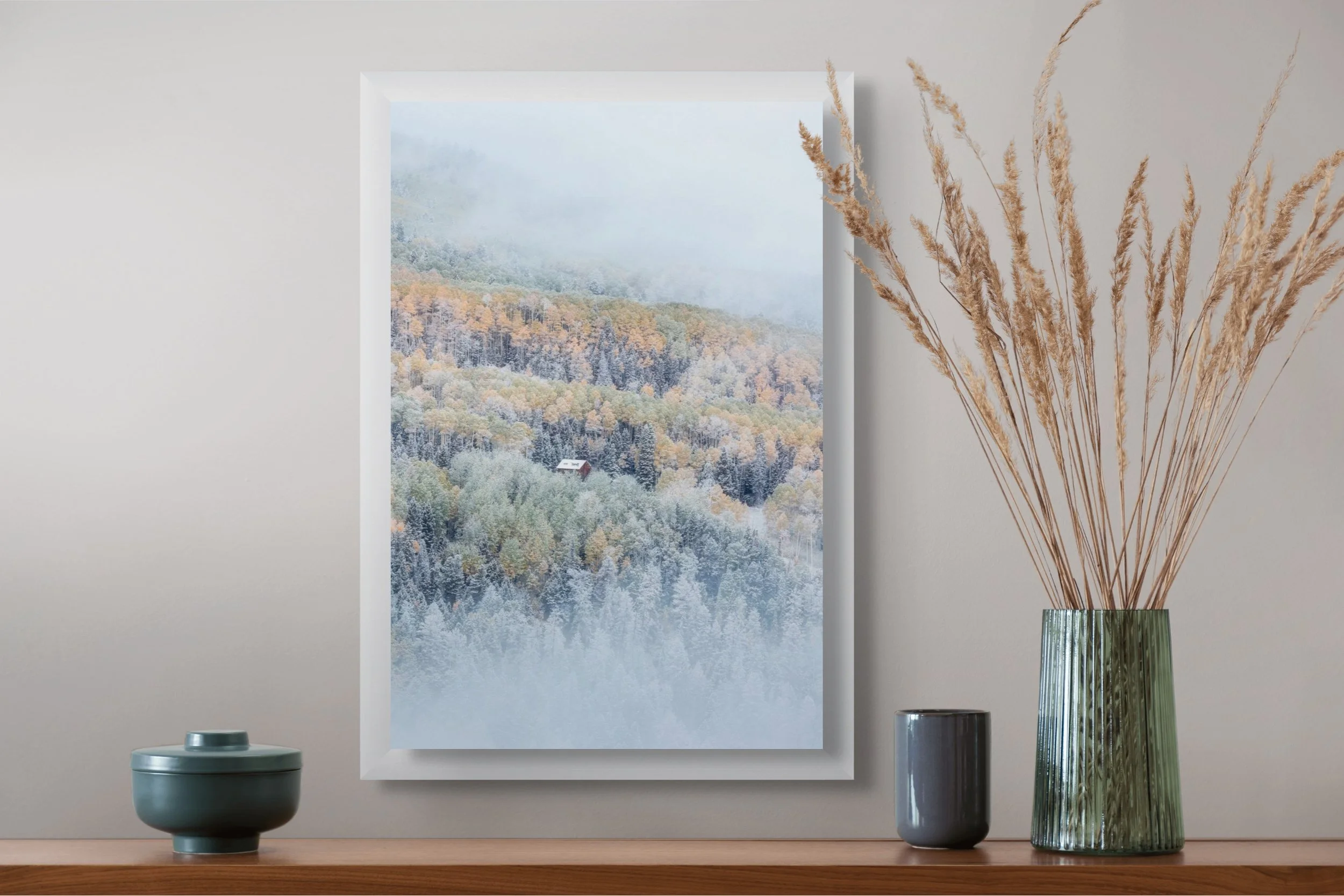 A framed landscape photograph of misty mountains and a forest with autumn foliage hangs on a light-colored wall above a wooden surface. On the surface, there are three vases: a small round blue-green vase with a lid, a taller dark gray cup, and a tal