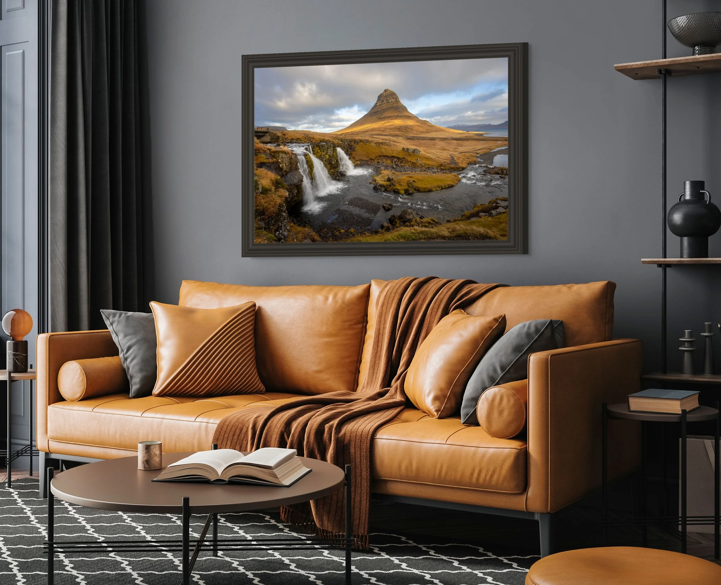 Living room with a tan leather sofa, pillows, a blanket, and a coffee table with an open book, under a wall art of a mountain landscape with waterfalls.