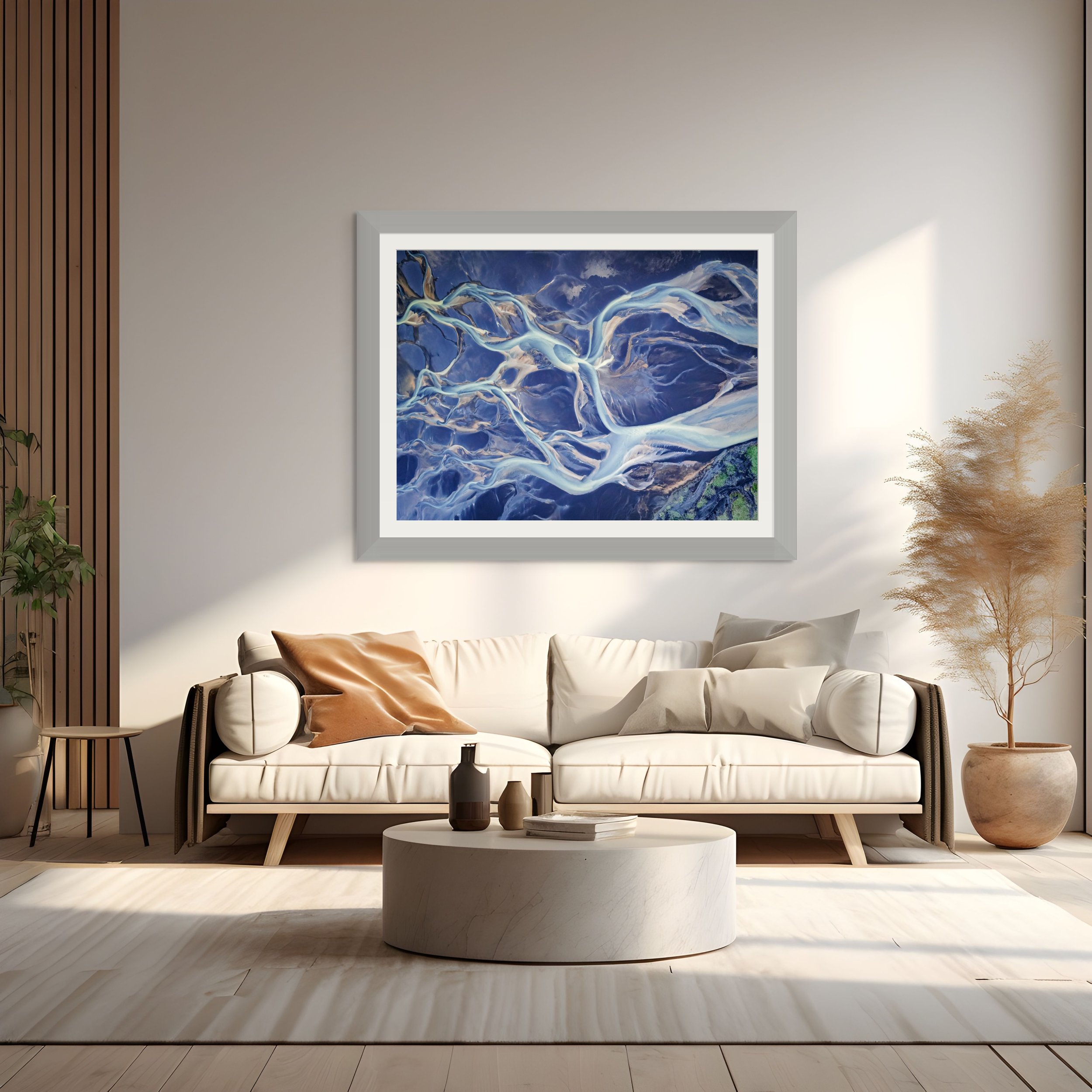 Modern living room with white sofa, beige and gray cushions, a round marble coffee table with vases and books, a wall with abstract blue and white artwork, light wood flooring, and a large potted plant in a beige pot.