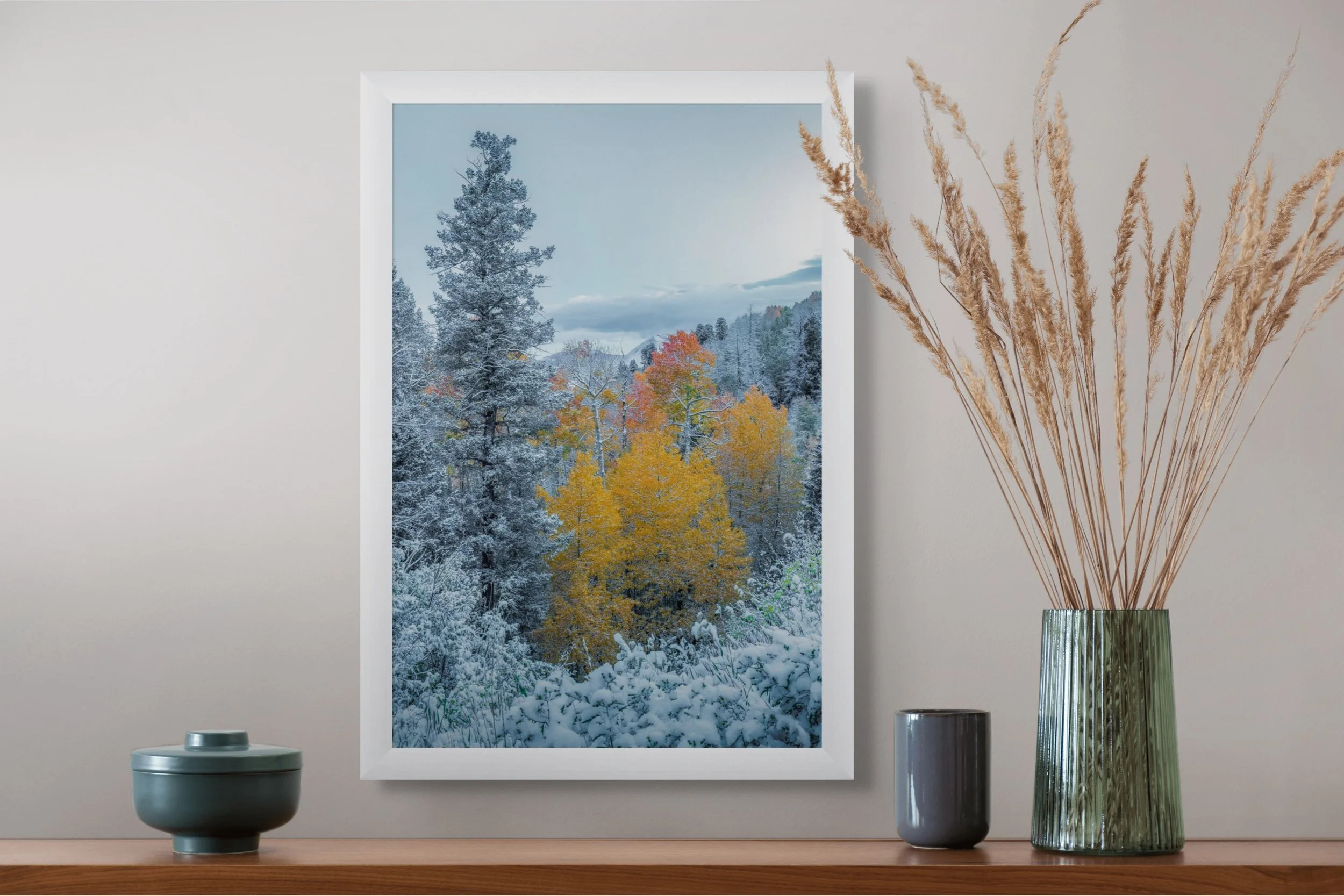 Interior scene with a framed winter landscape photo of snow-covered trees and colorful autumn leaves, placed on a wooden shelf next to a tall green vase with dried grass, a gray cup, and a small dark green container.