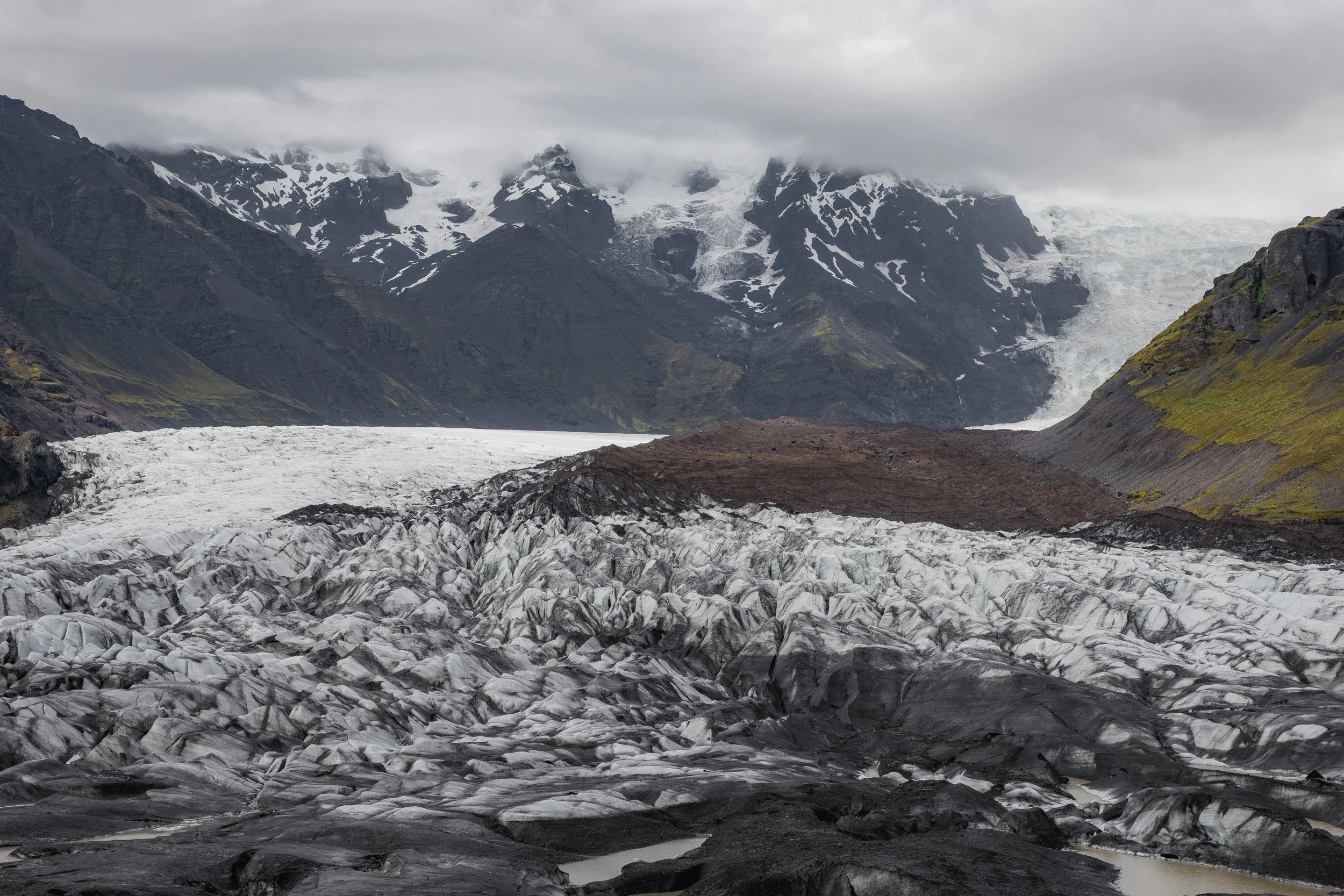 A dramatic view of an Icelandic glacier weaving between rugged volcanic peaks, where ancient ice meets raw earth. This powerful landscape captures the elemental contrast between fire-forged terrain and the timeless movement of ice.