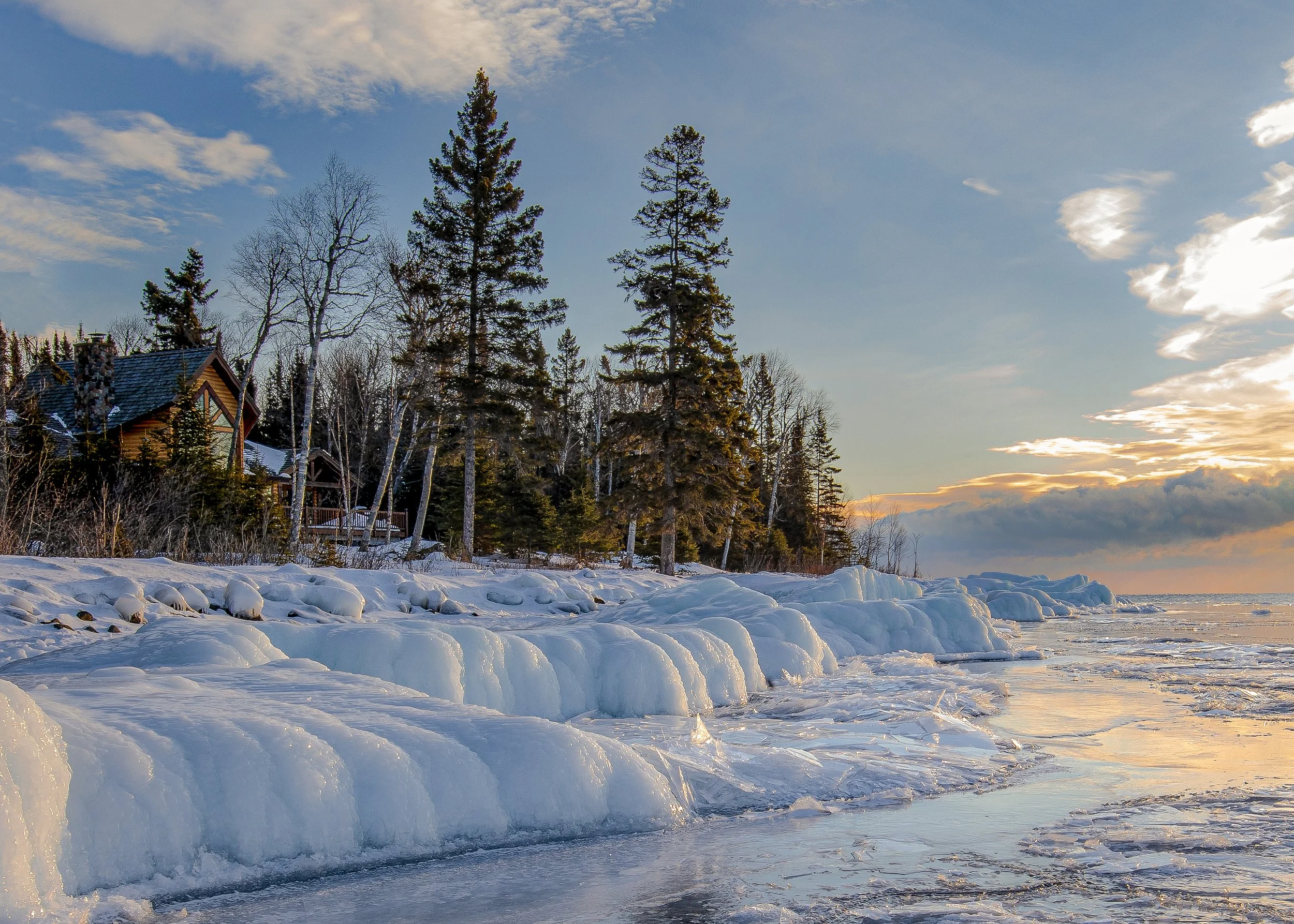 A rocky lakeshore covered in snow and wind-formed ice ridges runs along calm ice-covered water, with a wood and stone log cabin among bare birch trees and green spruce trees on the shore, under a blue sky with an orange sunset glow on the horizon.
