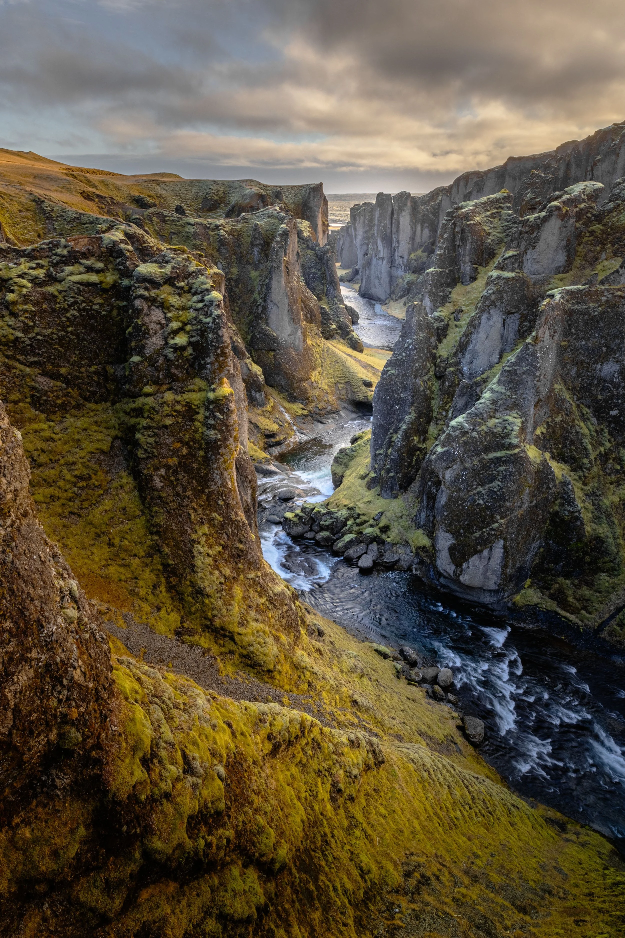 A rugged canyon with moss-covered rocks, a flowing river, and cloudy sky.