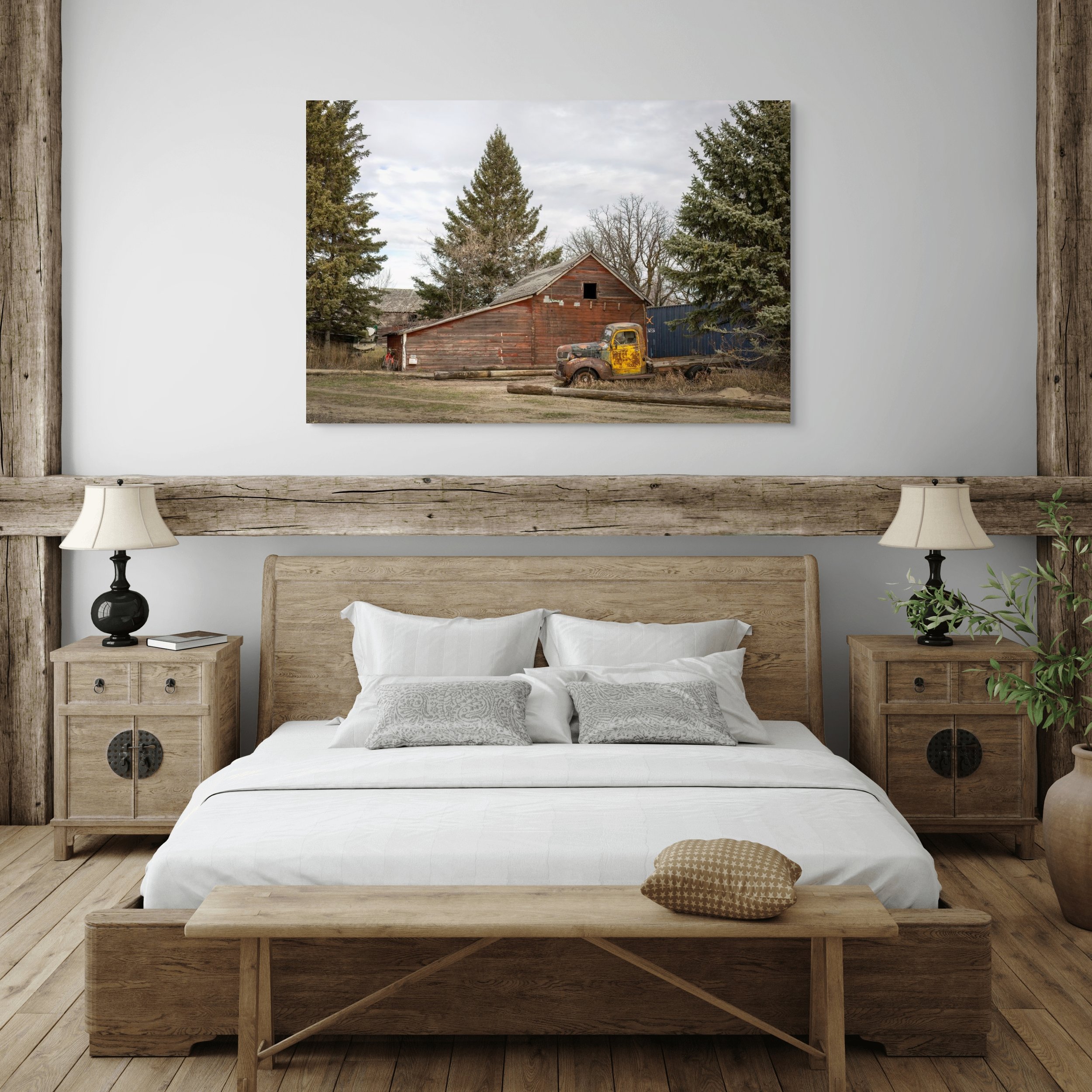 A bedroom with a wooden bed and matching nightstands, white bedding, gray pillows, two lamps, and a landscape painting of an old barn with trees and an abandoned yellow truck.