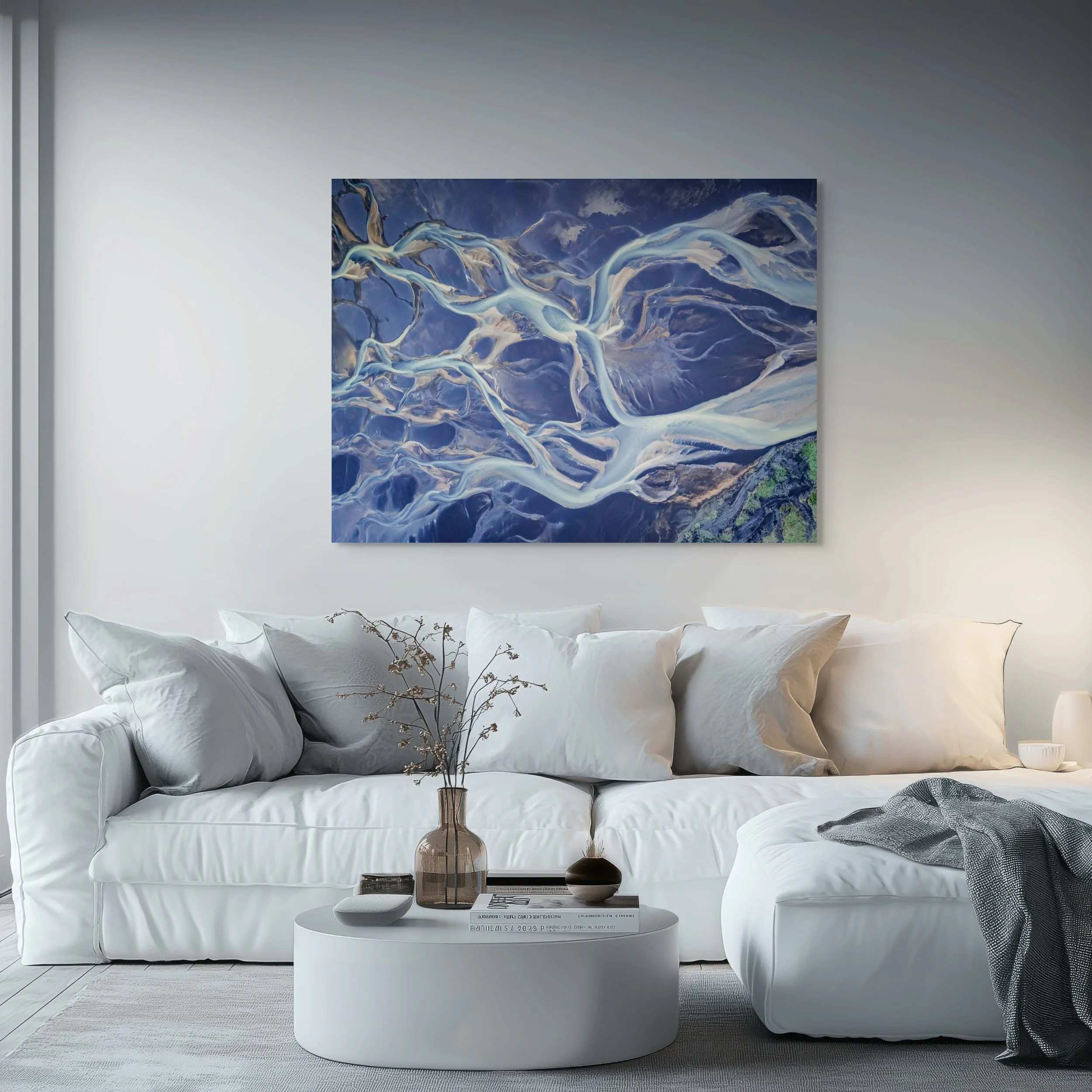 A living room with a white sofa, pillows, a small round table with a vase of dried flowers, and an abstract blue and white painting on the wall.
