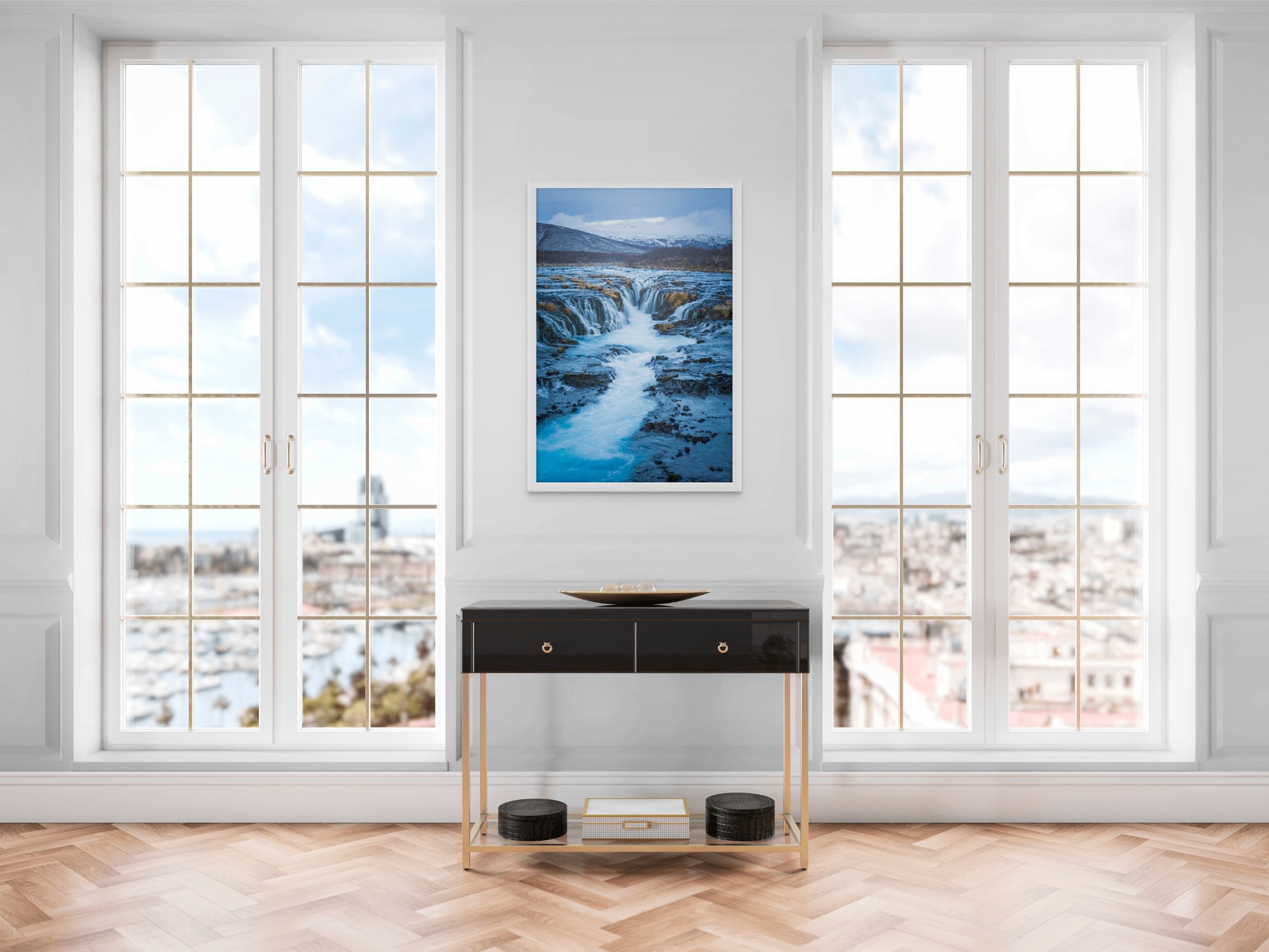Living room with large windows, a black console table, a framed landscape photograph of a river and mountains, and a view of city buildings in the background.
