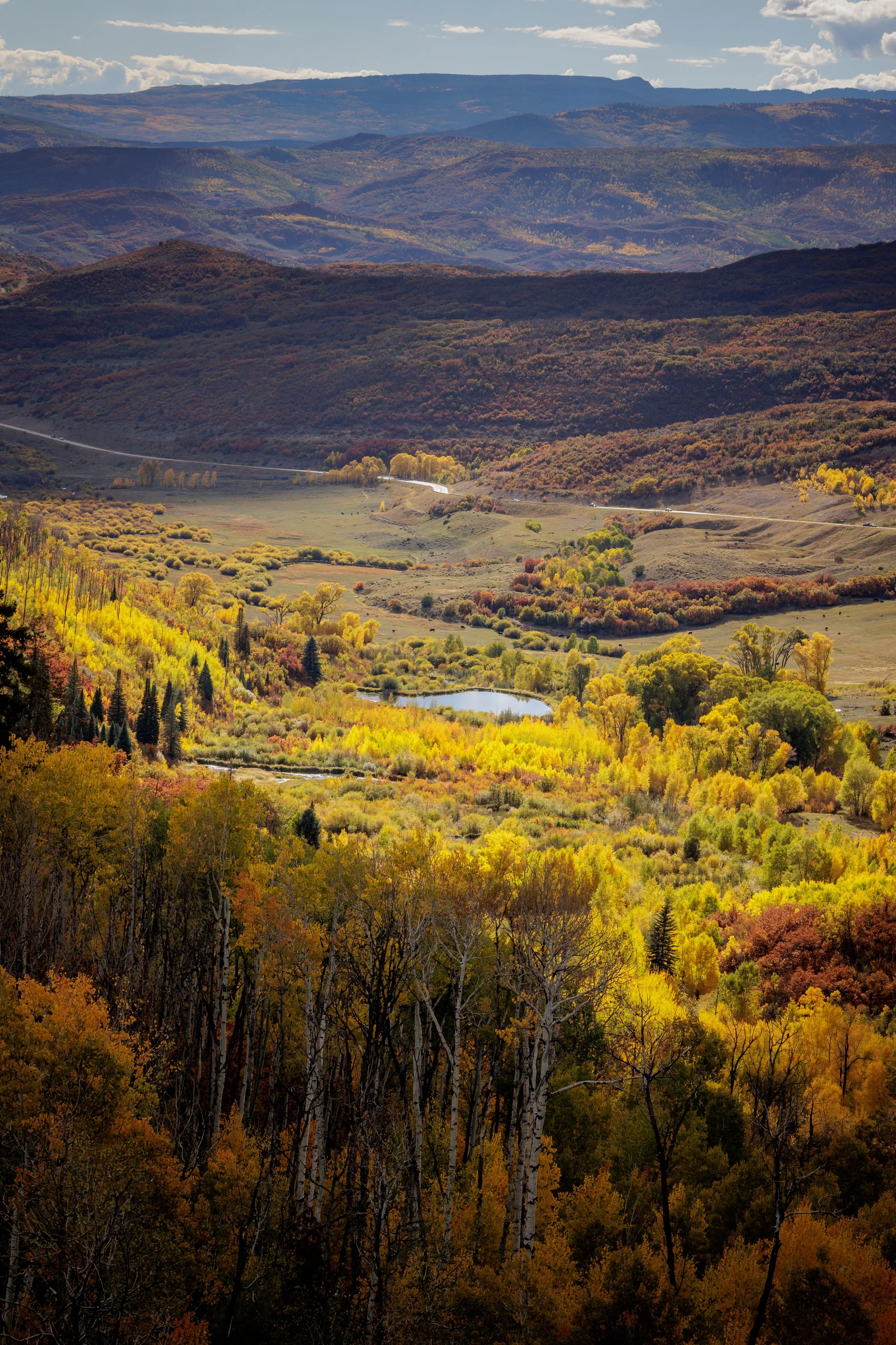 A scenic landscape of rolling hills and mountains covered in colorful autumn trees, with a small pond and a winding road visible in the valley.