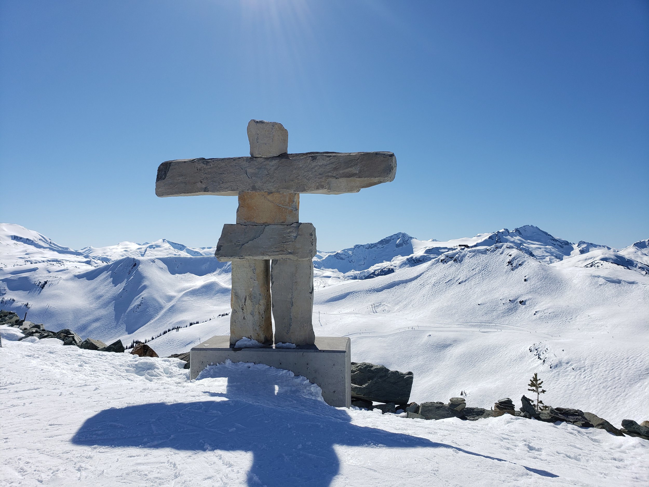 A stone cross monument on a snowy mountain with snow-covered peaks and blue sky in the background.