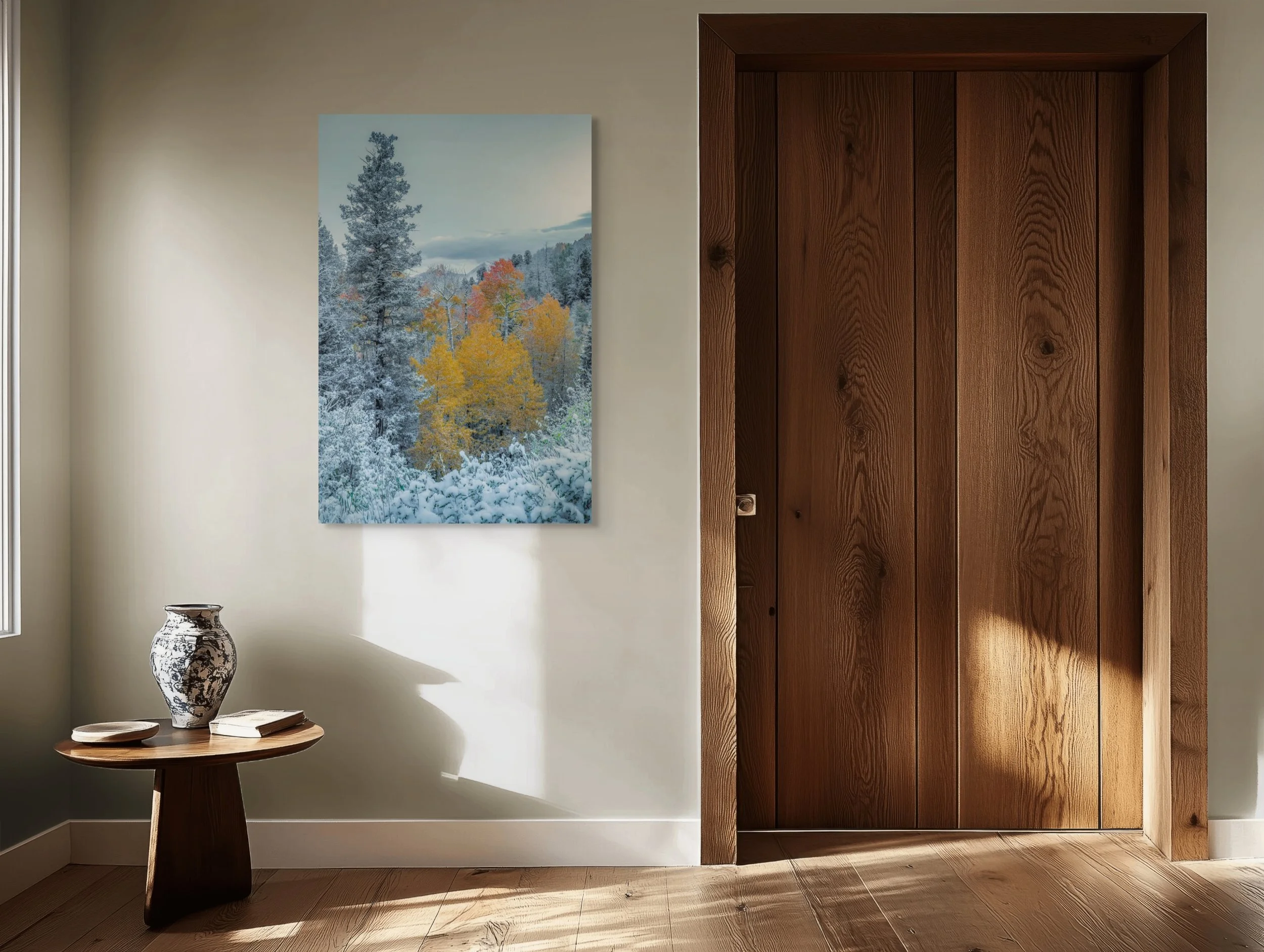 A room with a wooden door, a framed landscape photo of snow-covered trees and colorful fall trees on the wall, a round wooden side table with a black and white vase, a book, and a small bowl, and sunlight casting shadows on the wall and floor.