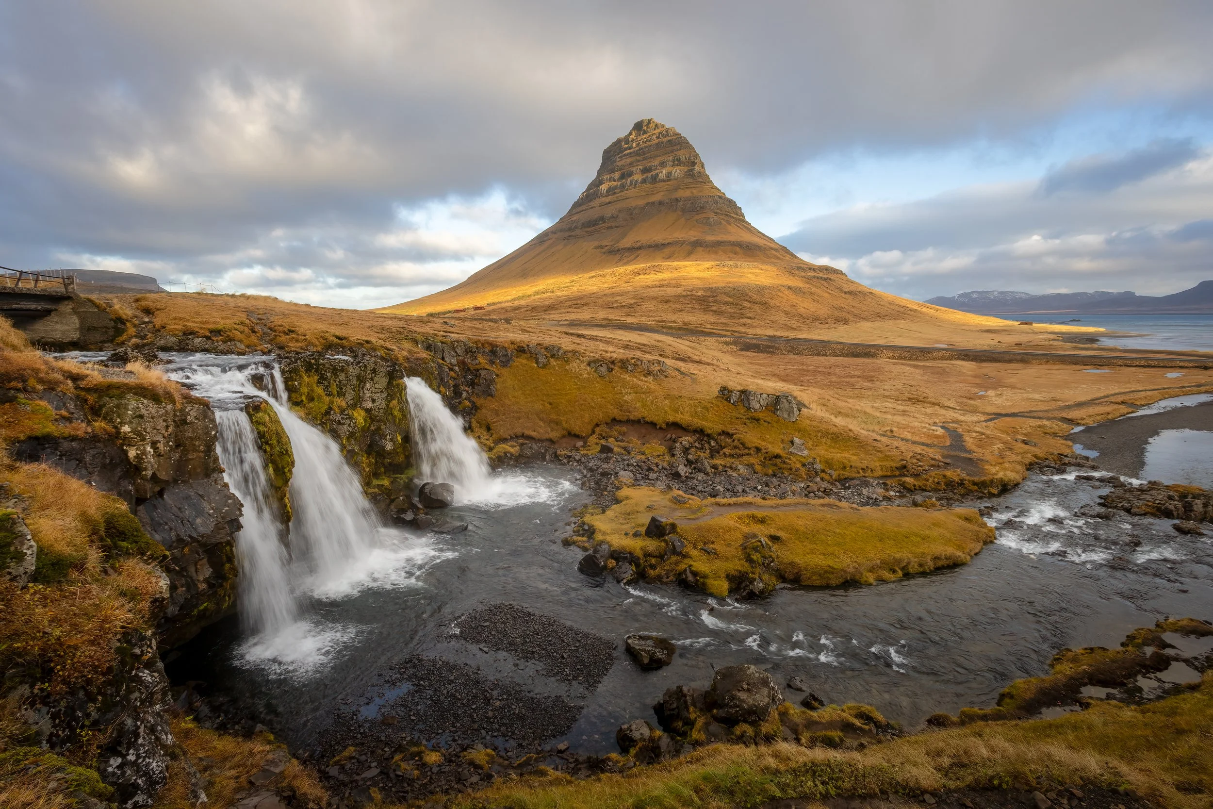 A scenic landscape featuring a conical mountain in the background, a small waterfall in the foreground, and a river flowing from the waterfall across grassy terrain under a cloudy sky.