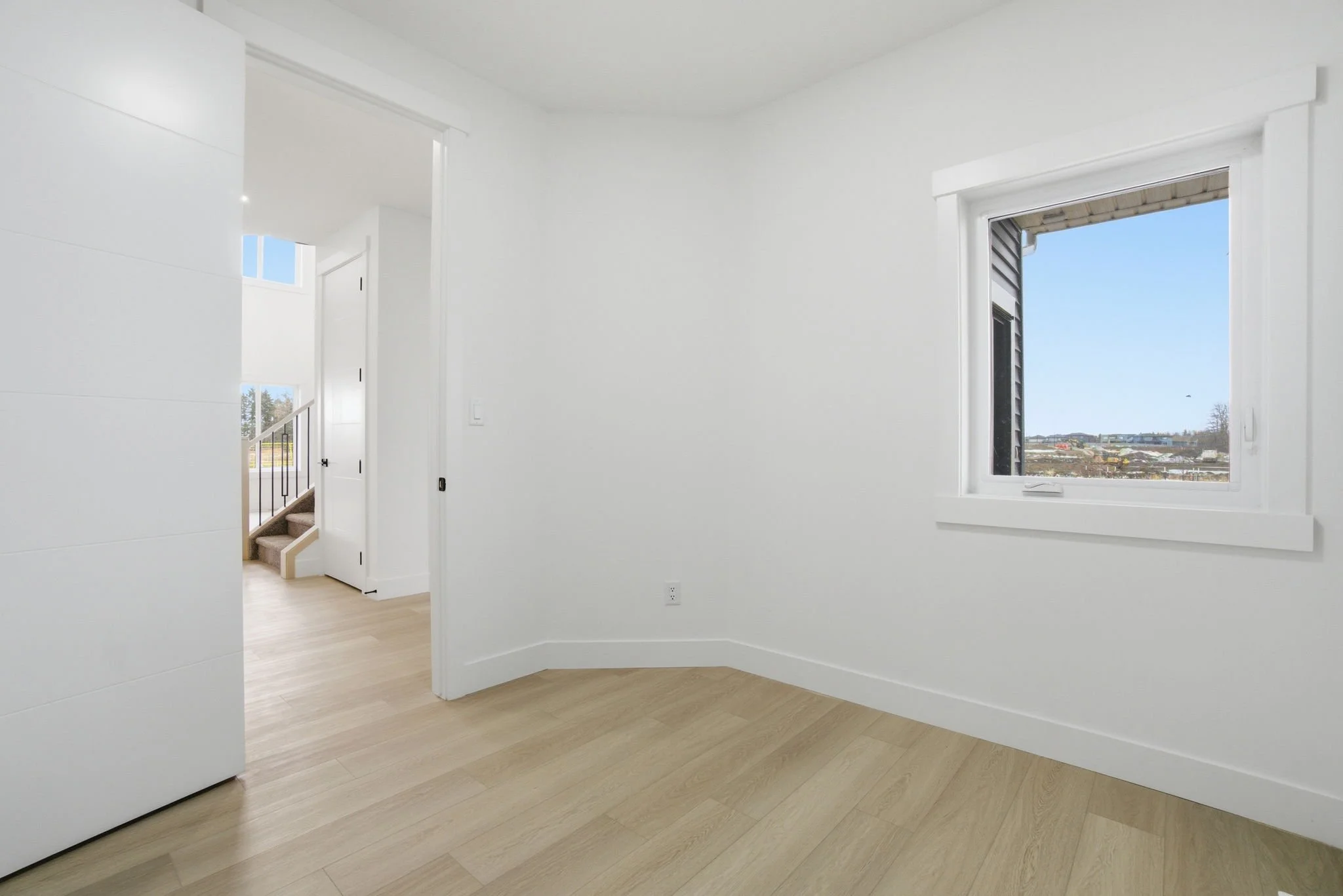 Interior view of a bright, white-walled room with large window showing a clear sky and outdoor landscape, light wood flooring, and a staircase leading upstairs.