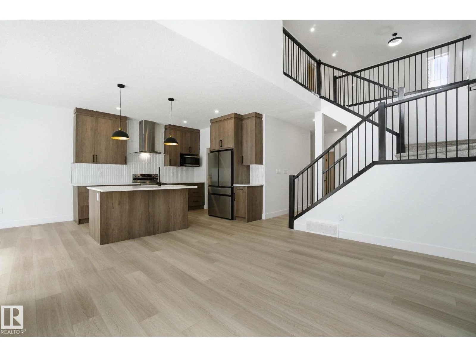 Open-concept kitchen with wood cabinets, stainless steel refrigerator, island with a white countertop, and black pendant lights, adjacent to a staircase with black railing.
