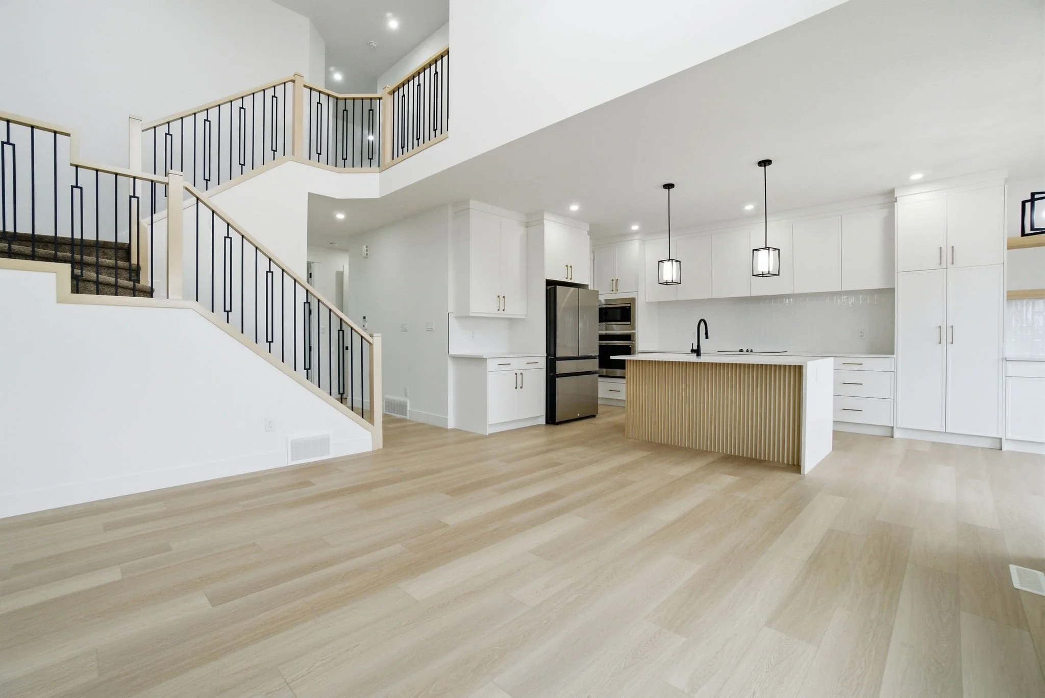 A modern open-concept kitchen and living area with white cabinetry, stainless steel appliances, a kitchen island with a light wood vertical slat design, pendant lighting, light wood flooring, and a staircase with black and light wood railing.