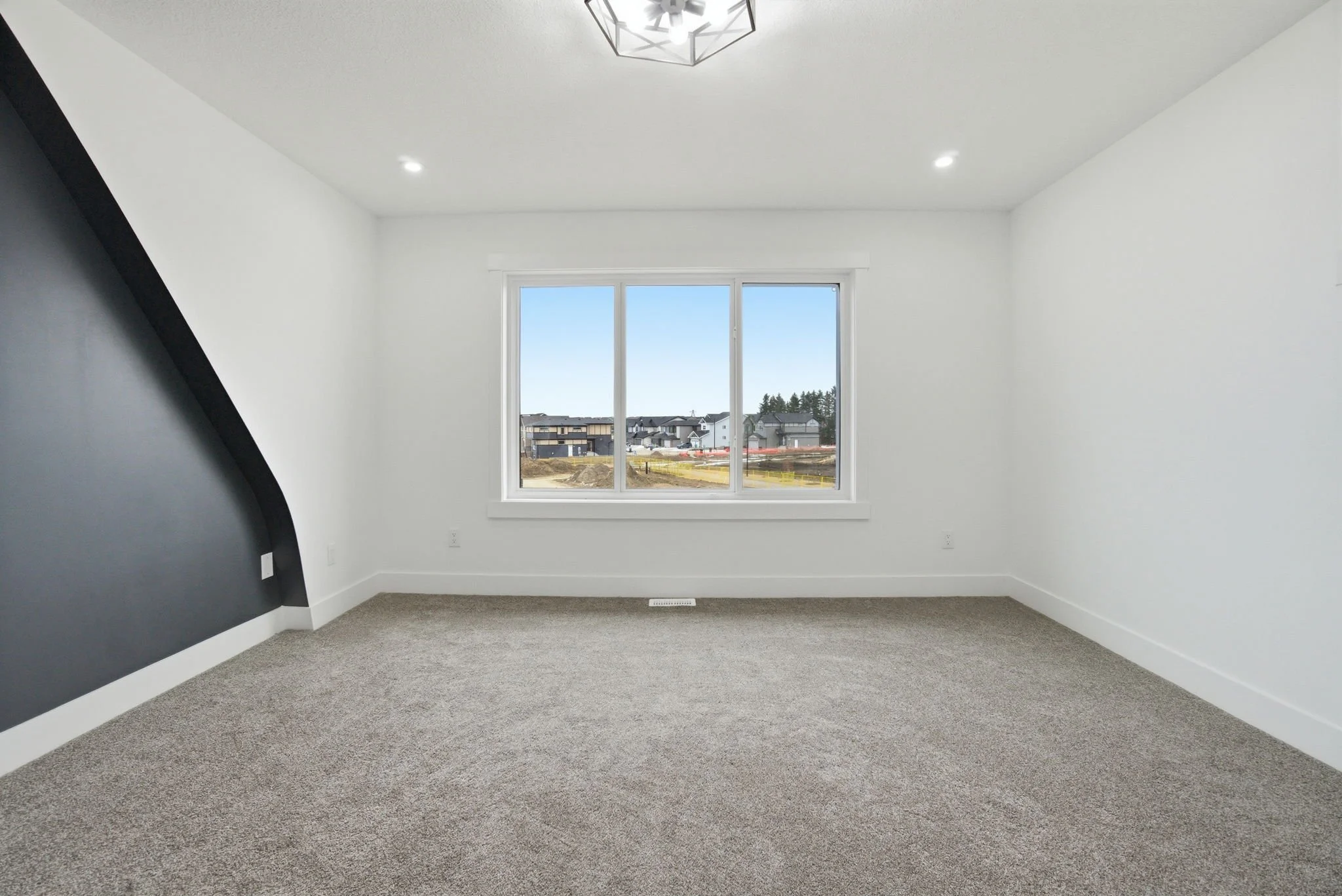 Empty room with a large window showing a suburban neighborhood outside. The room has white walls, a gray accent wall, gray carpet, and modern ceiling lights.