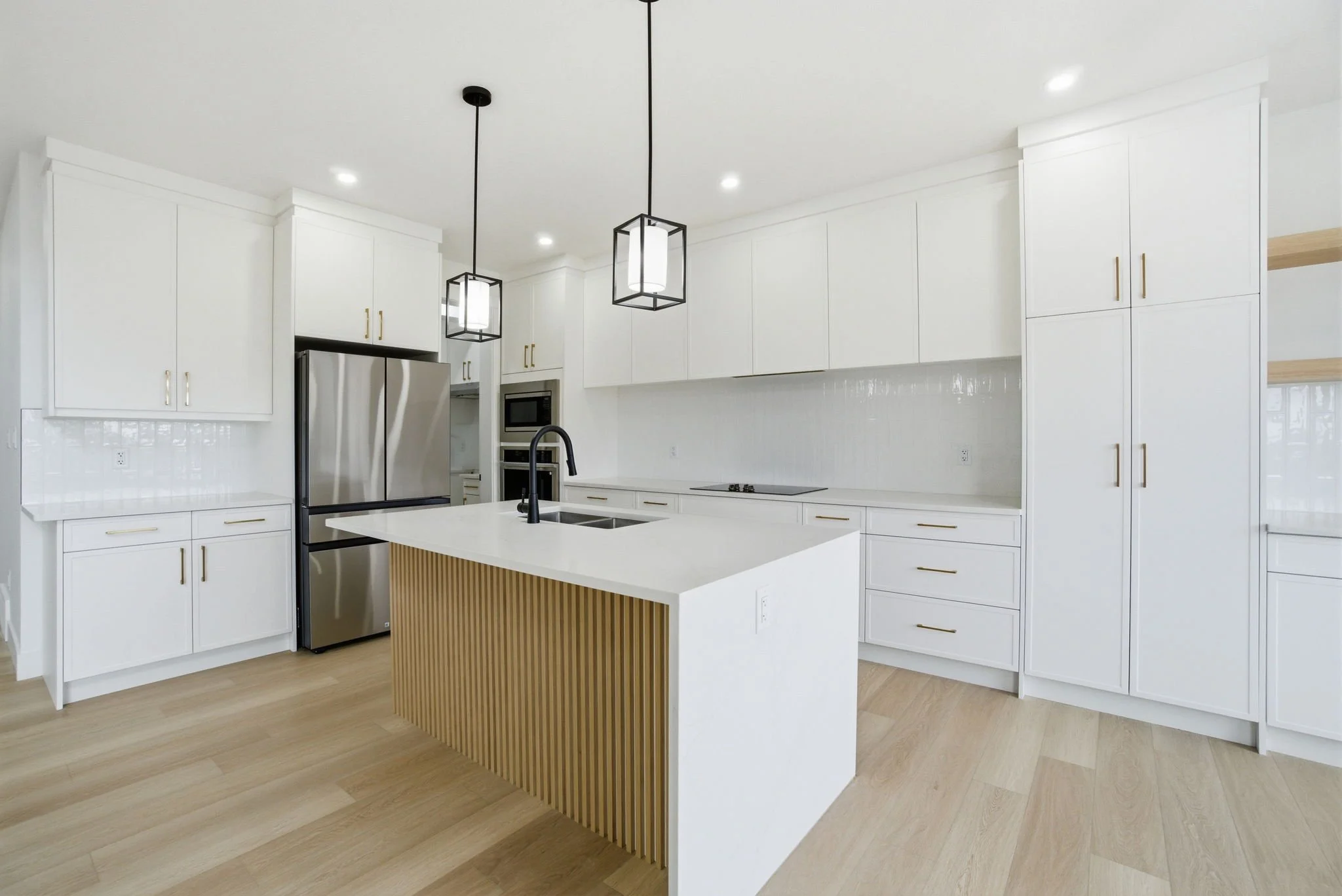Modern white kitchen with wooden accents, island with black faucet, stainless steel refrigerator, built-in microwave and oven, pendant and recessed lighting, light wood flooring.