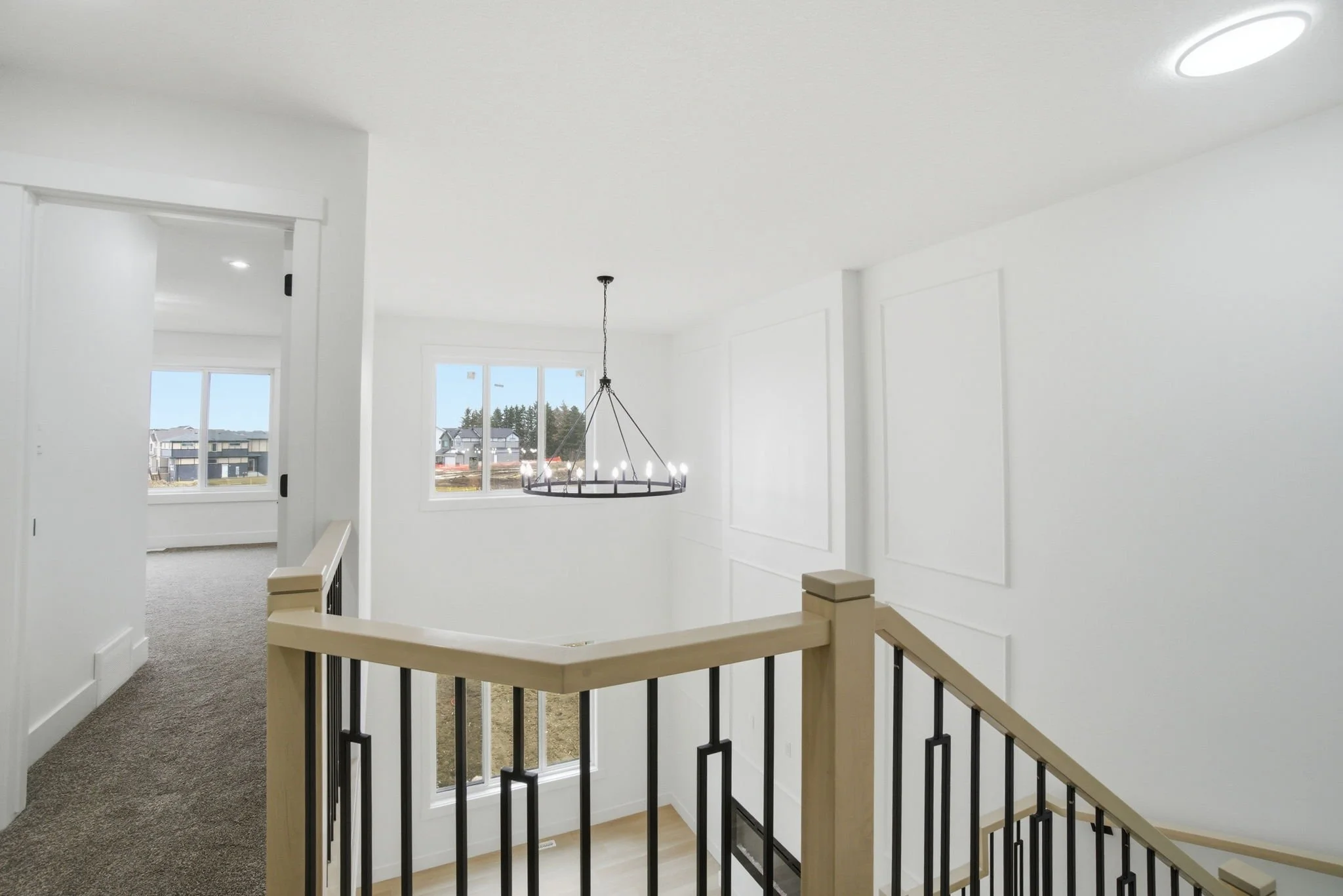 Interior view of a modern home with white walls, a chandelier, large windows, and an open staircase with a wooden and metal railing.